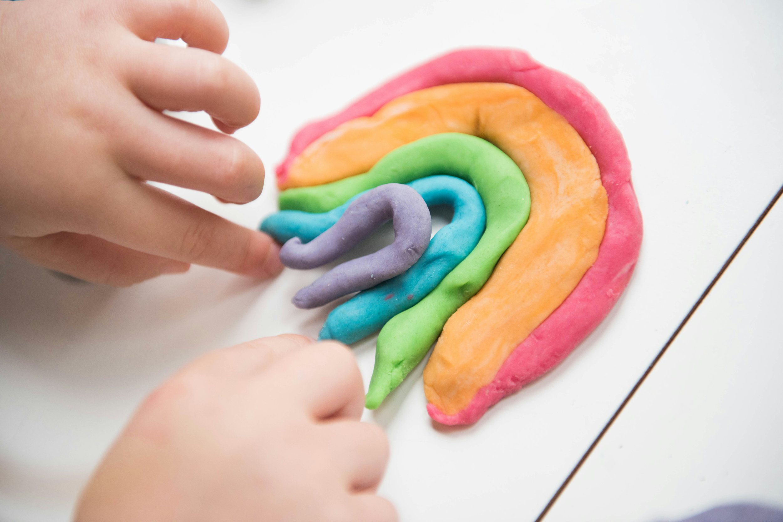Child's hand decorating a rainbow-shaped cookie with colorful icing on a white surface.