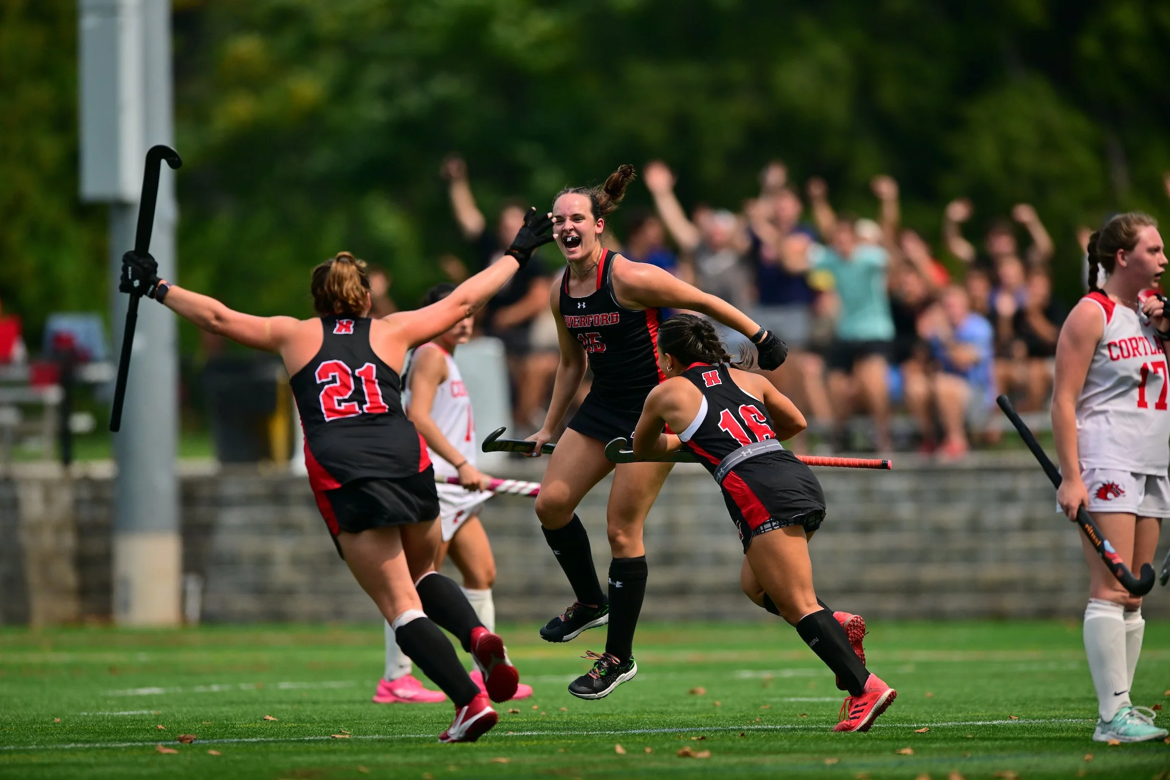 A group of Haverford field hockey players celebrating on the field during the win over nationally-ranked SUNY Cortland. The background shows spectators watching the game.