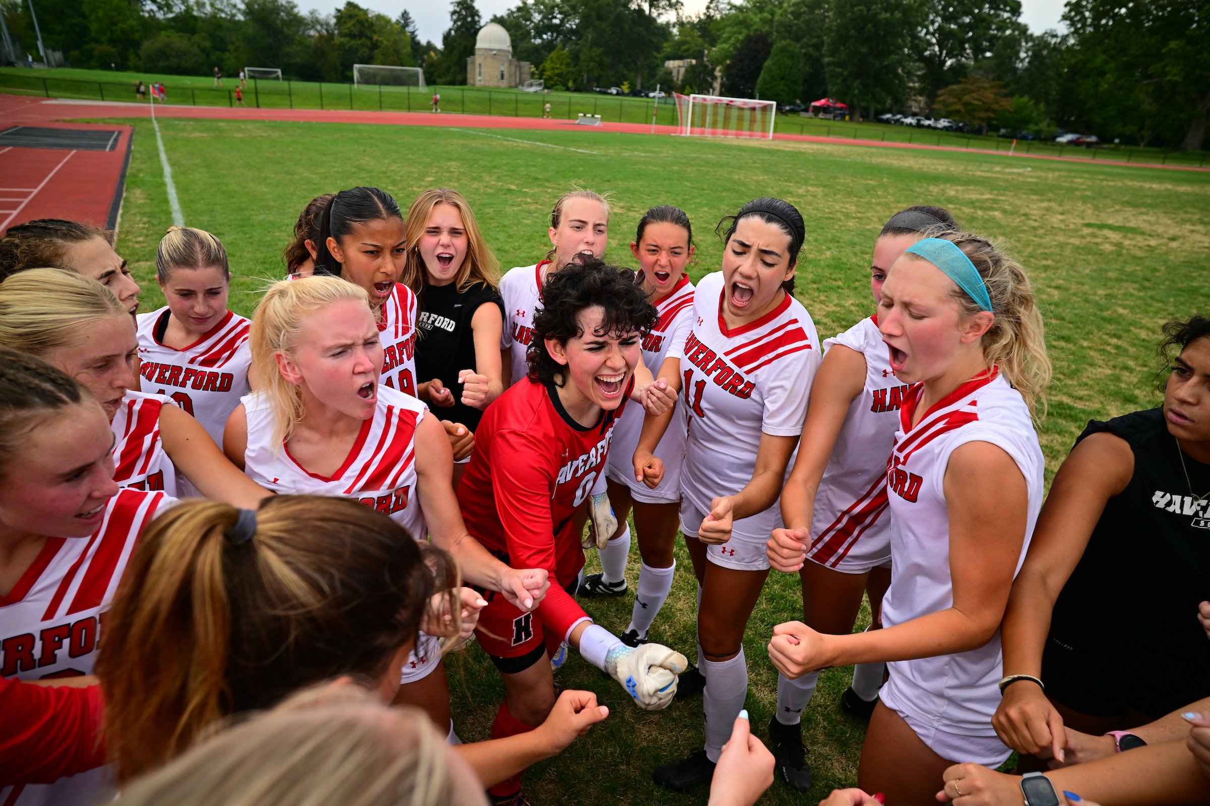 Group of Haverford women's soccer players in white uniforms with red lettering huddle and cheer before a home contest. The background shows a running track, goalposts, and Haverford's observatory building.