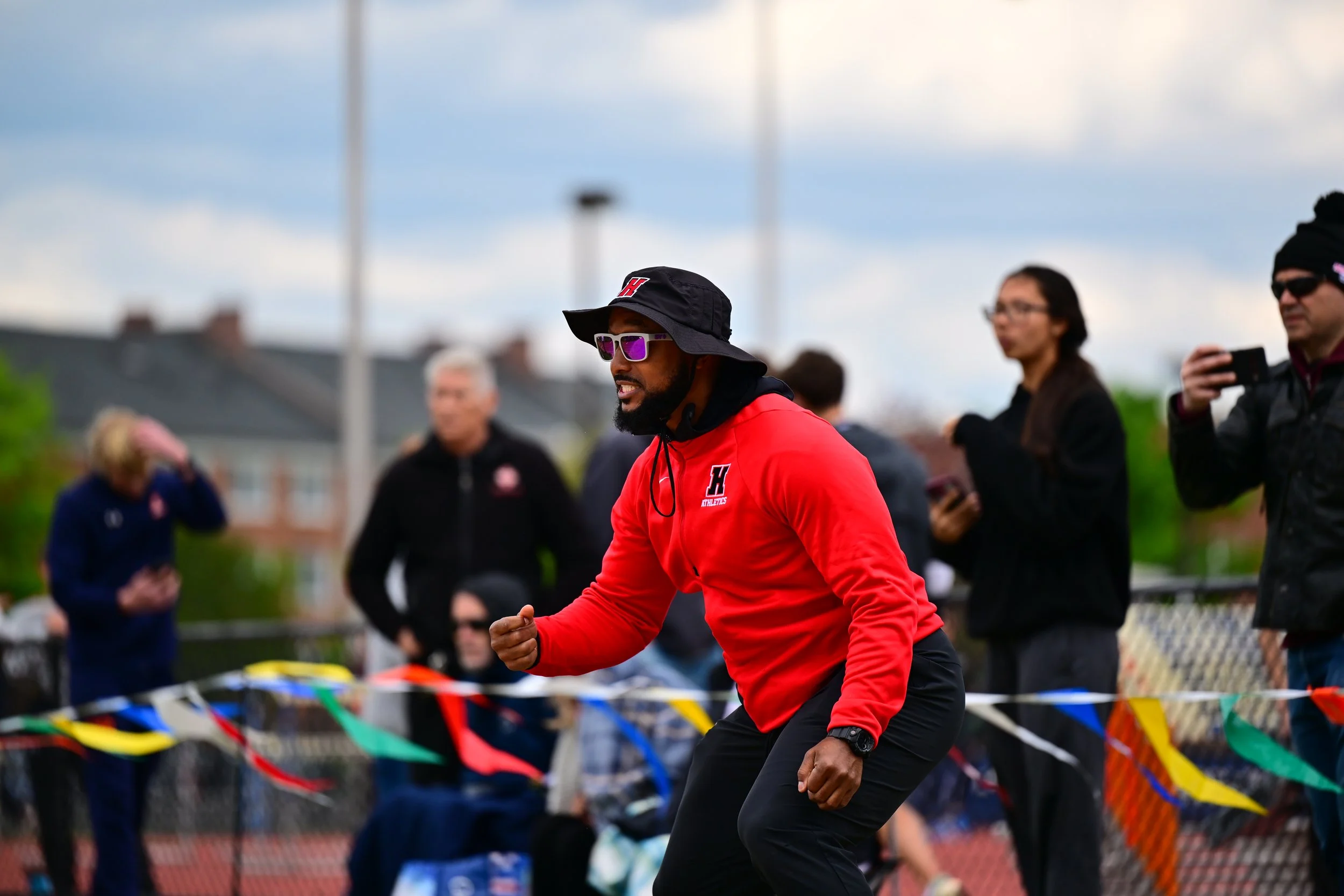 A man in a red jacket and black hat with a logo, wearing sunglasses, is running on a track during an outdoor event, with a few spectators and colorful flags in the background.