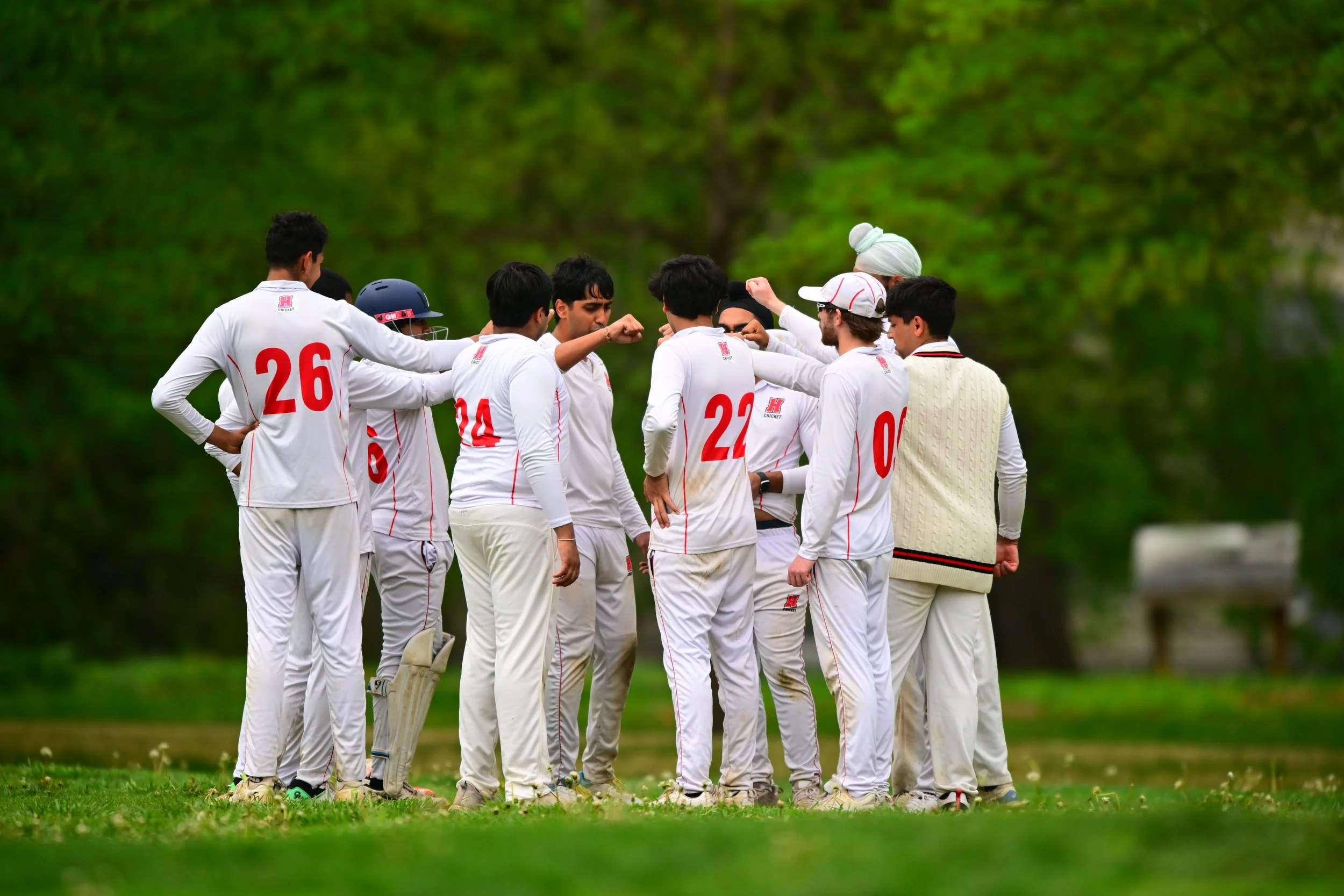 The Haverford cricket team in white uniforms, huddled together on a grassy field, preparing for play, with the arboretum campus in the background.