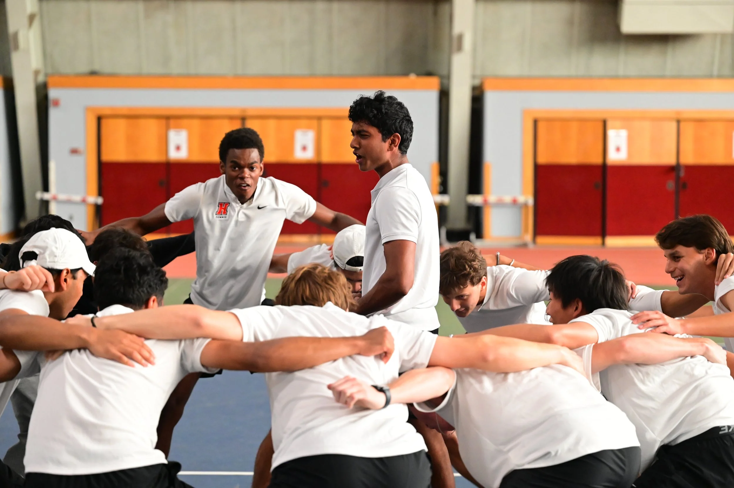 Group of students in athletic uniforms participating in a team huddle or cheer in an indoor gym or sports facility.