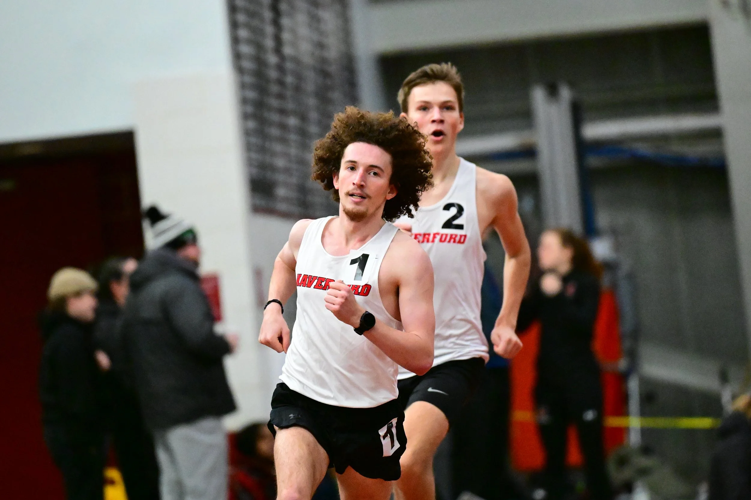 Two Haverford Men's Track & Field athletes participating in indoor track race, wearing Haverford uniforms with lane numbers, running at a fast pace.