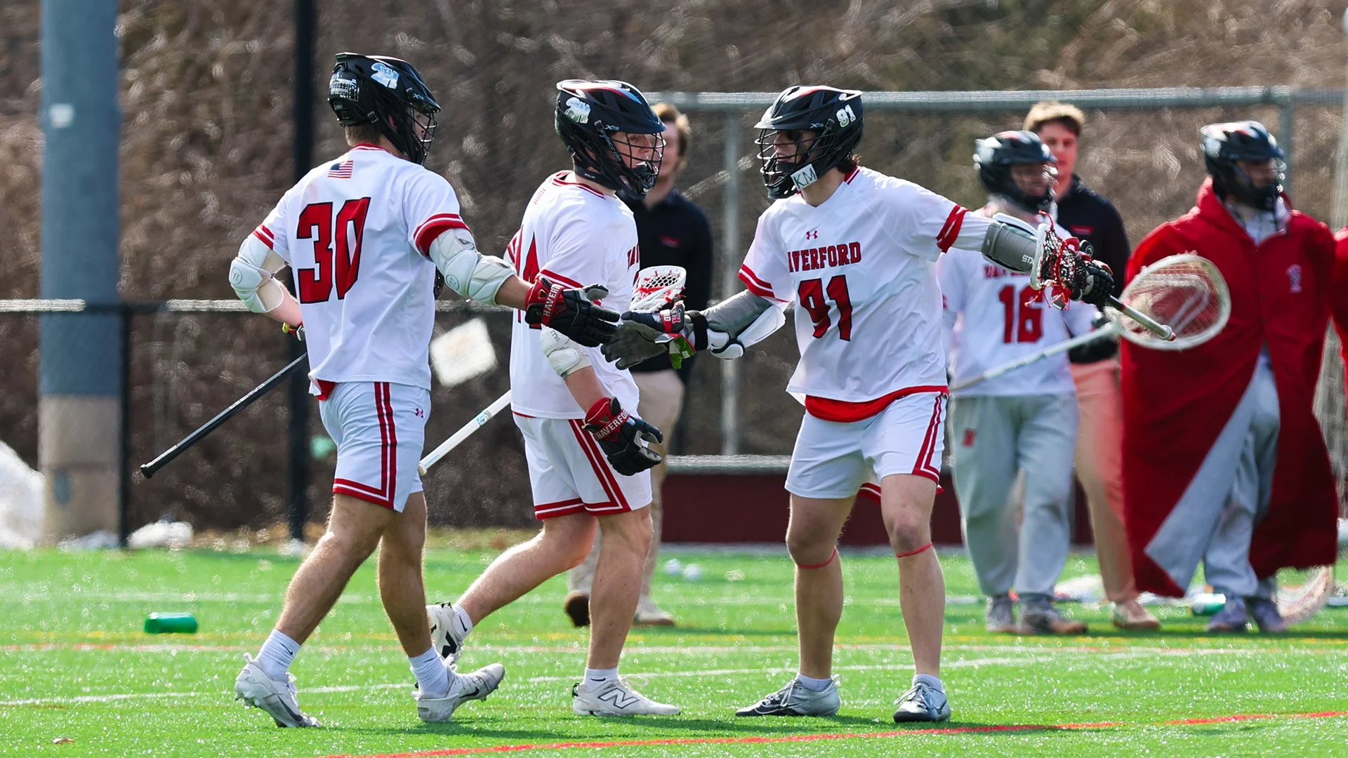 Three lacrosse players in white uniforms celebrate with a handshake on the field while teammates and coaches watch from the sideline.