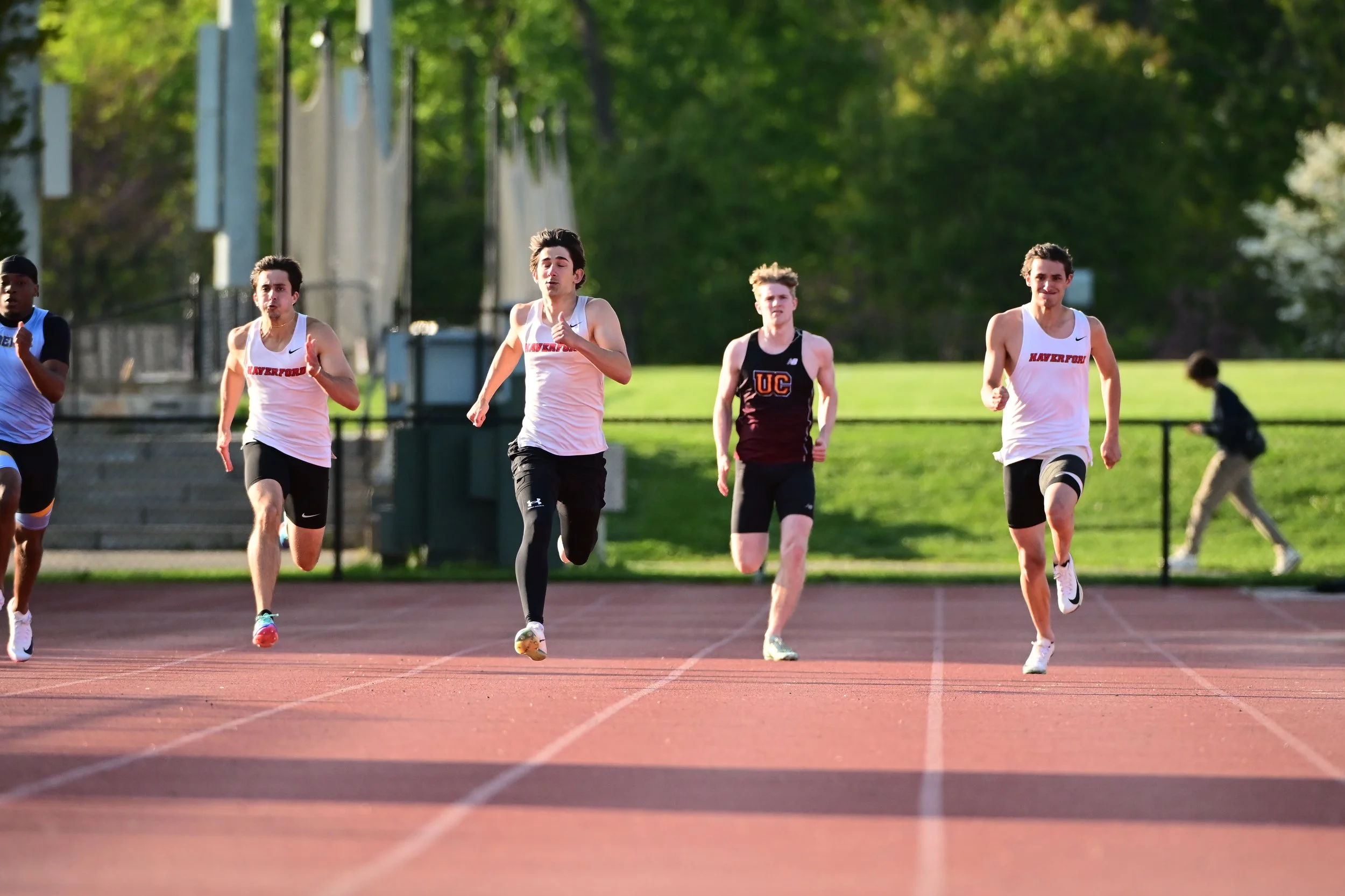 A group of young male runners participating in a track race outdoors, with trees and a fence in the background.
