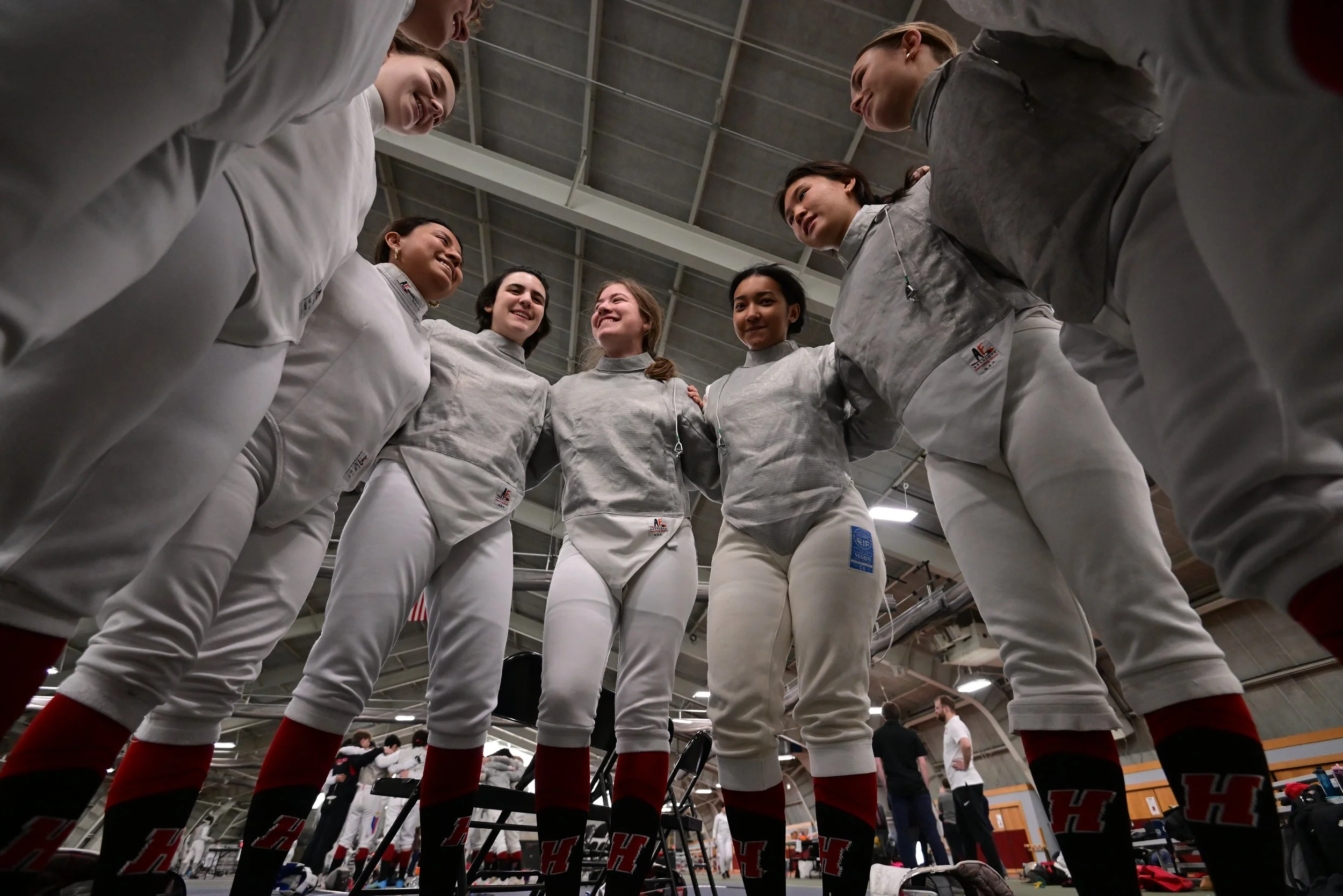 A group of Haverford women's fencing team members standing in a circle with their arms around each other's shoulders, smiling and talking, inside Alumni Field House.