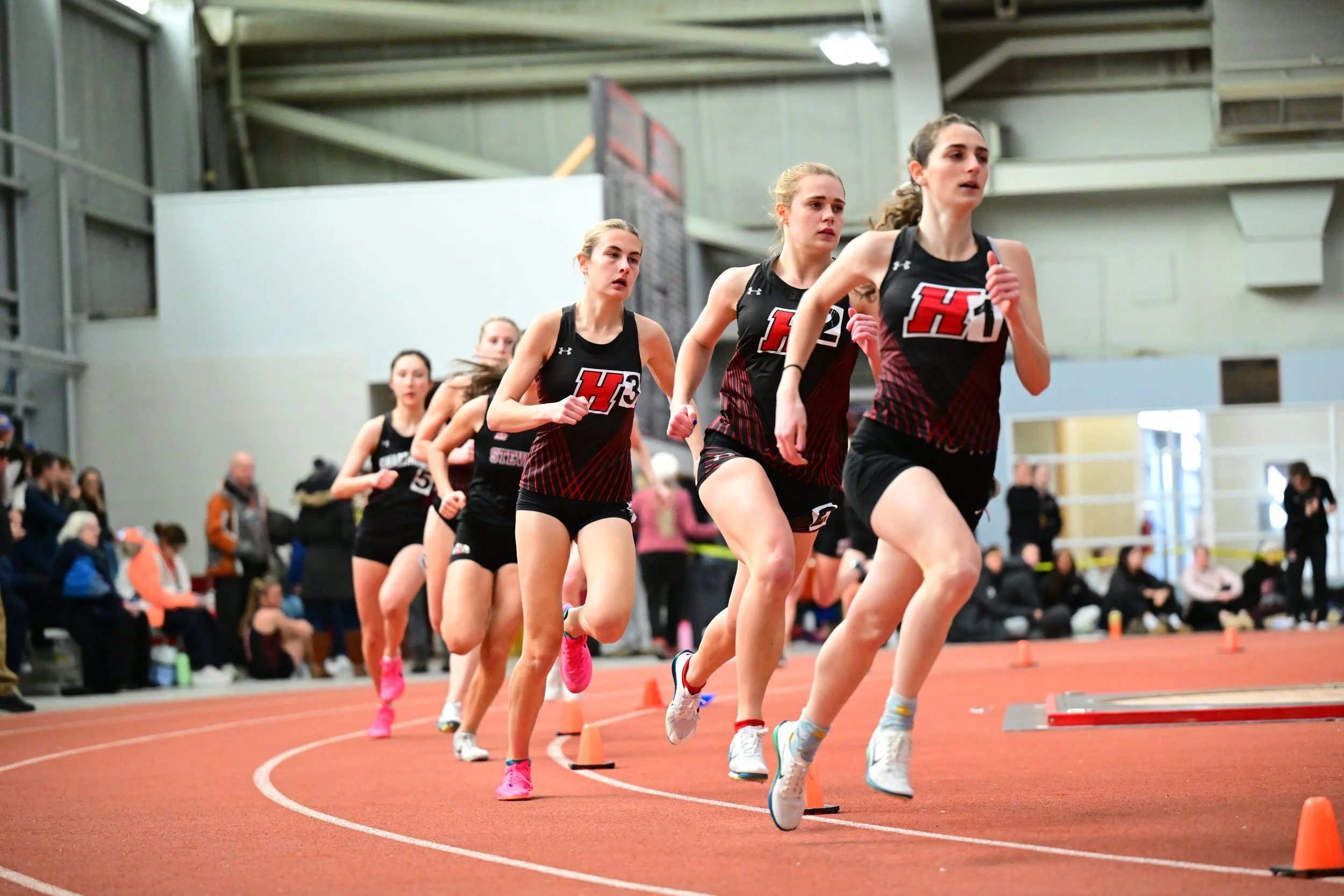 Several Haverford Women's Track & Field athletes participating in an indoor track race, wearing Haverford uniforms with lane numbers, running at a fast pace.