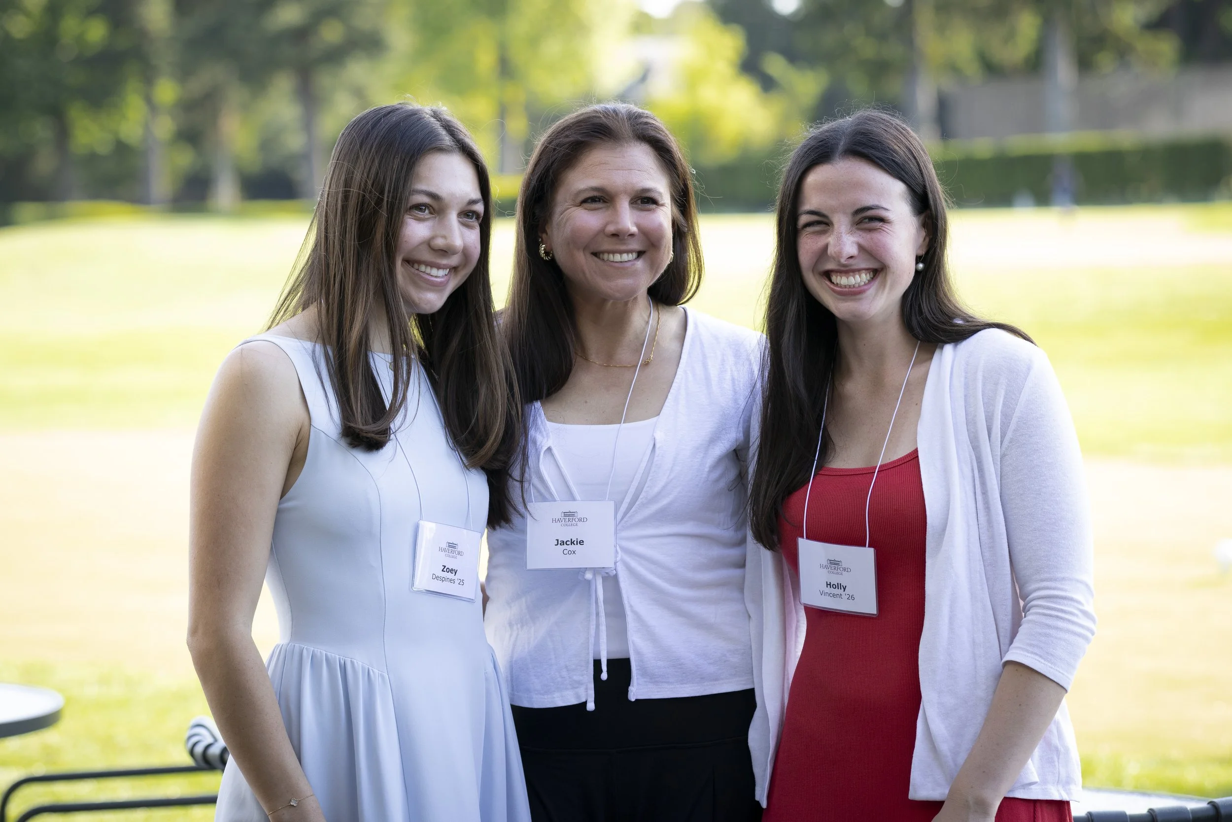 Three women standing outdoors at an event, smiling, wearing name tags, with trees and grass in the background.