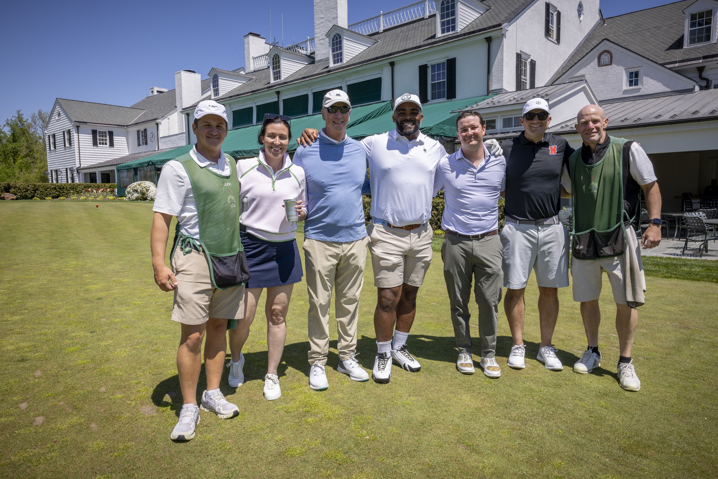 Group of seven people standing on a golf course, smiling, with a large white house in the background under a clear blue sky.