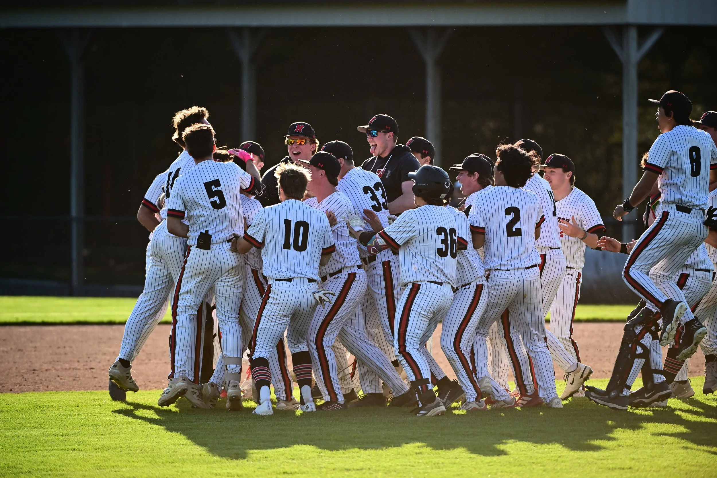 A group of Haverford baseball players celebrating a victory on Kannerstein Field, many wearing pinstriped uniforms with numbers, some wearing sunglasses, with the outfield fence and Haverford's arboretum campus trees in the background.