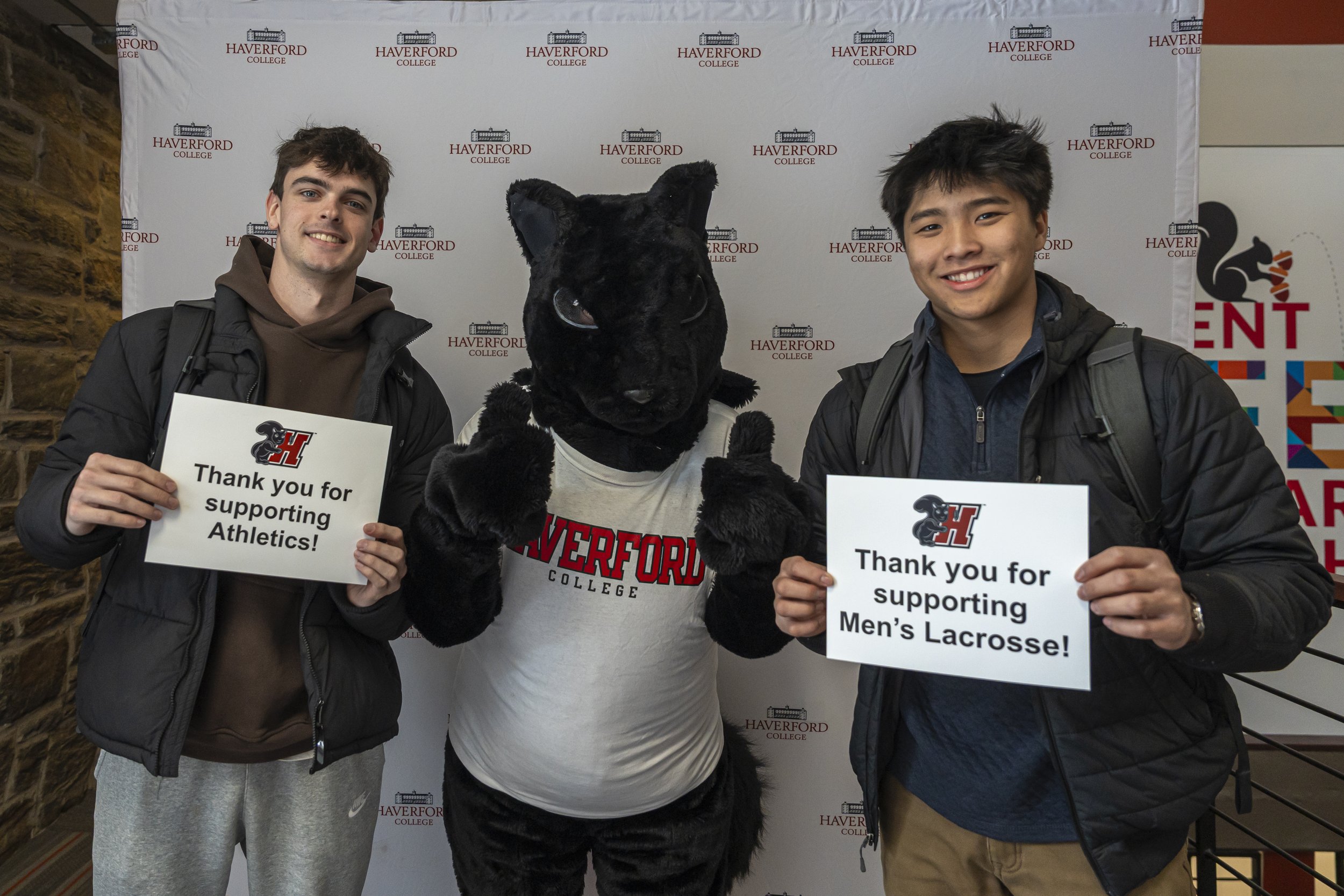 Two young men and a person in a black panther mascot costume are standing in front of a white background with the Haverford College logo. The two men are holding signs that say 'Thank you for supporting Athletics!' and 'Thank you for supporting Men's Lacrosse!' They are smiling and wearing backpacks.