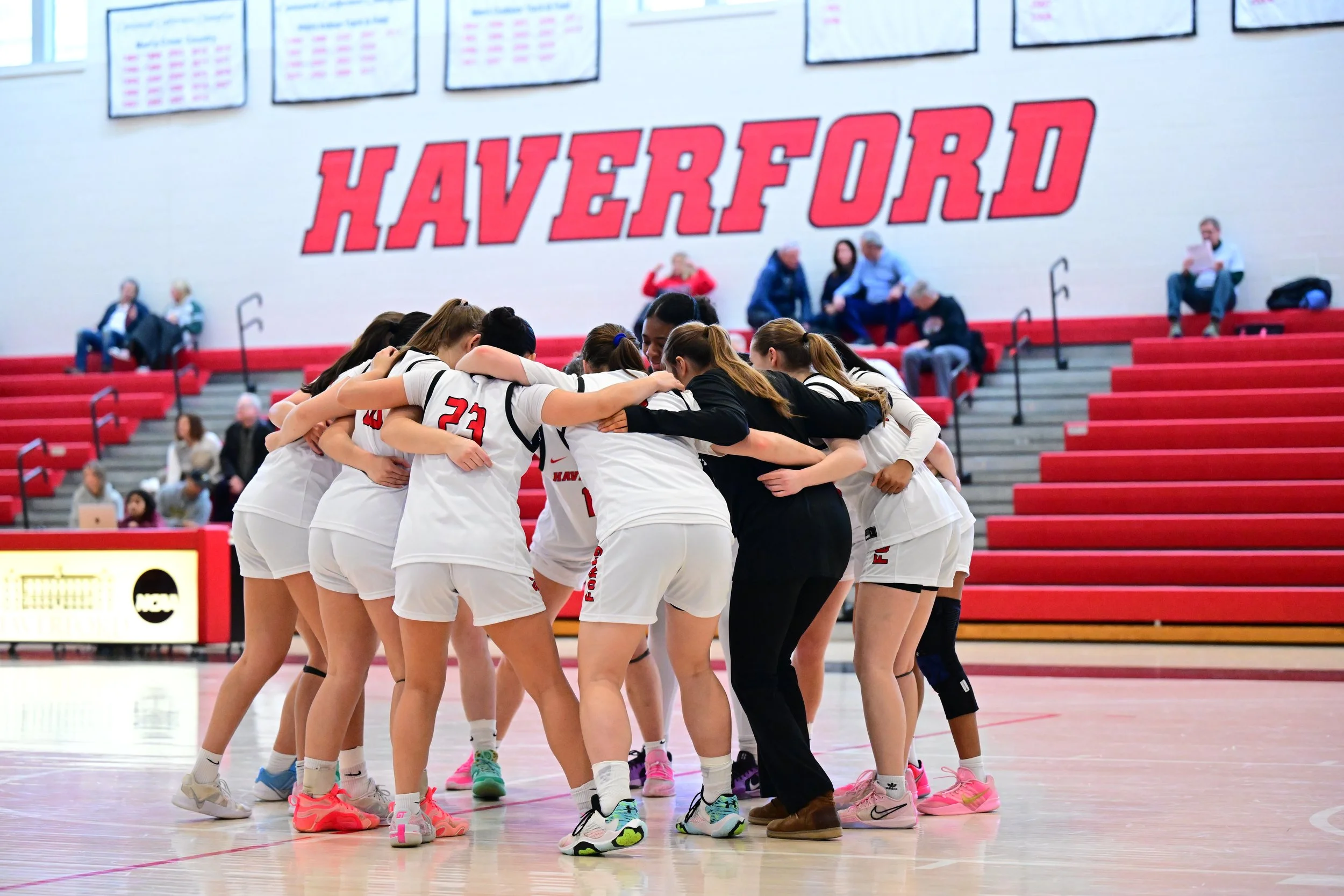 A group of Haverford Women's Basketball players in white uniforms huddle together. Behind them, a large red sign reads 'Haverford' on the wall, with spectators seated on red bleachers and some people sitting at the top of the bleachers.