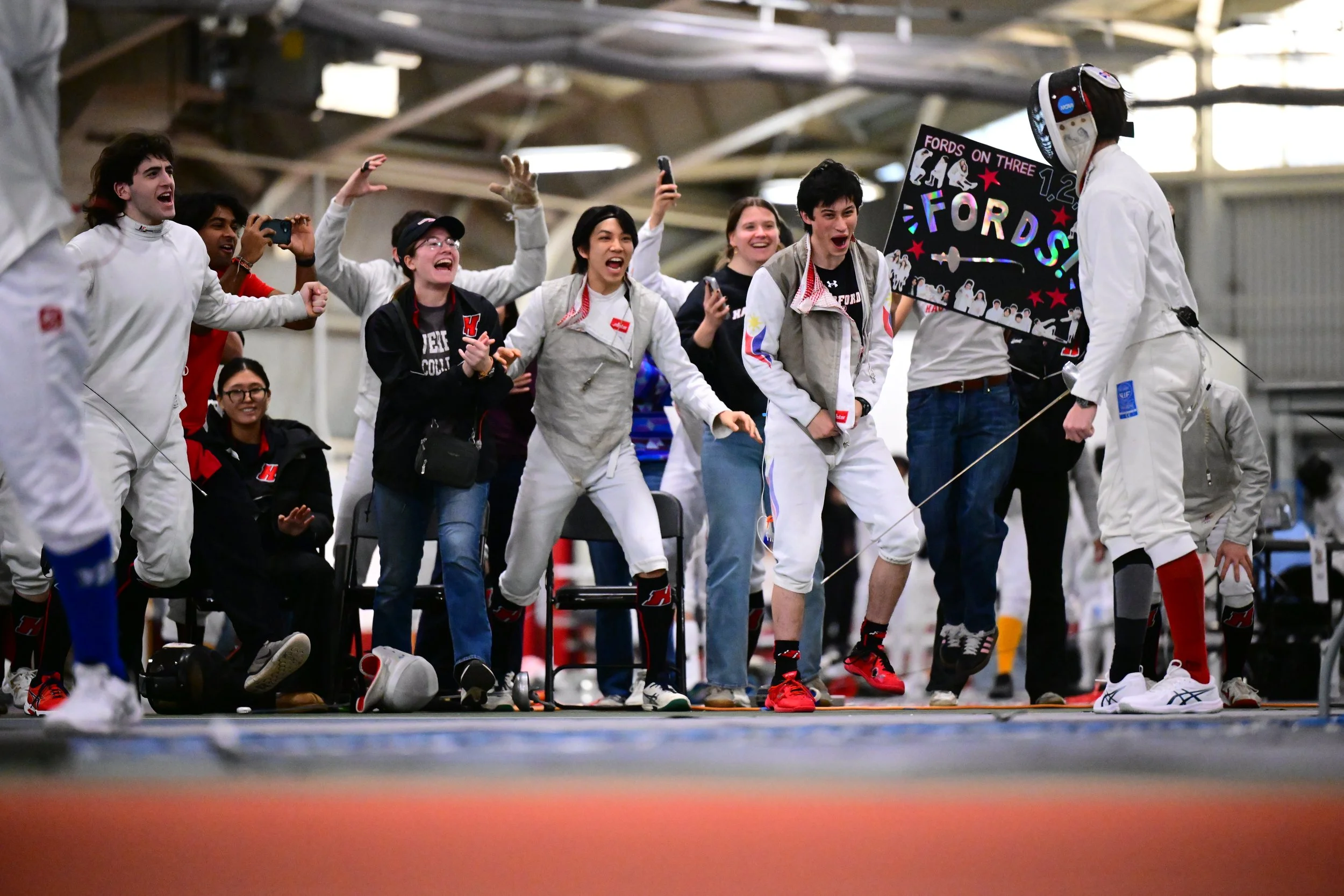 A group of Haverford fencers, most of whom are wearing their fencing gear, celebrating indoors at Alumni Field House. One person is holding a sign that reads 'Fords on Three' with a star, while others are taking photos and smiling.