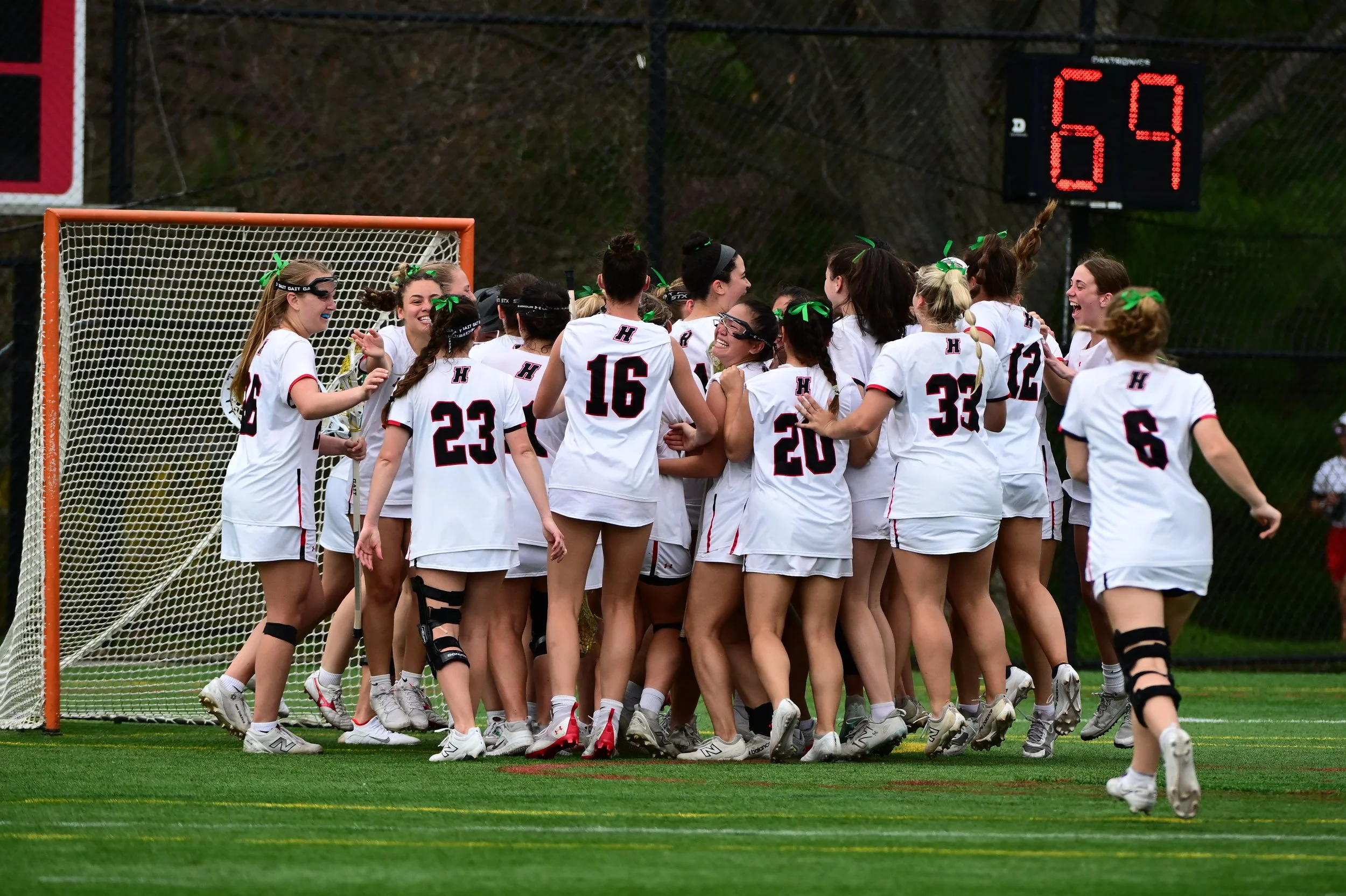 A group of Haverford Women's Lacrosse players in white jerseys celebrating on Swan Field near the goal, with a digital scoreboard showing the time remaining on the shot clock at the time the game ended.