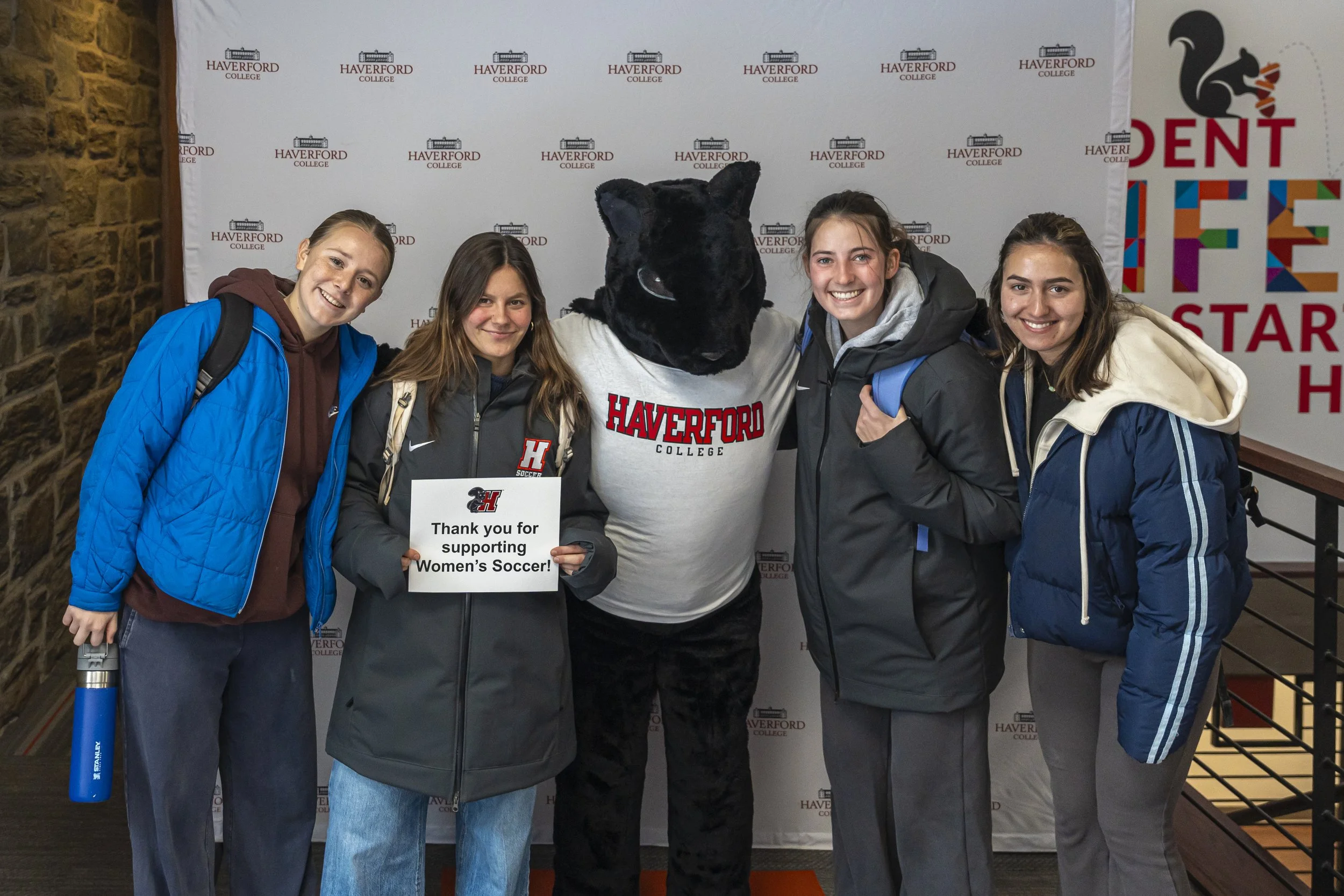 Four young women with backpacks standing with a person in a black cat costume at Haverford College, holding a sign about supporting women's soccer, in front of a backdrop with Haverford College logos.