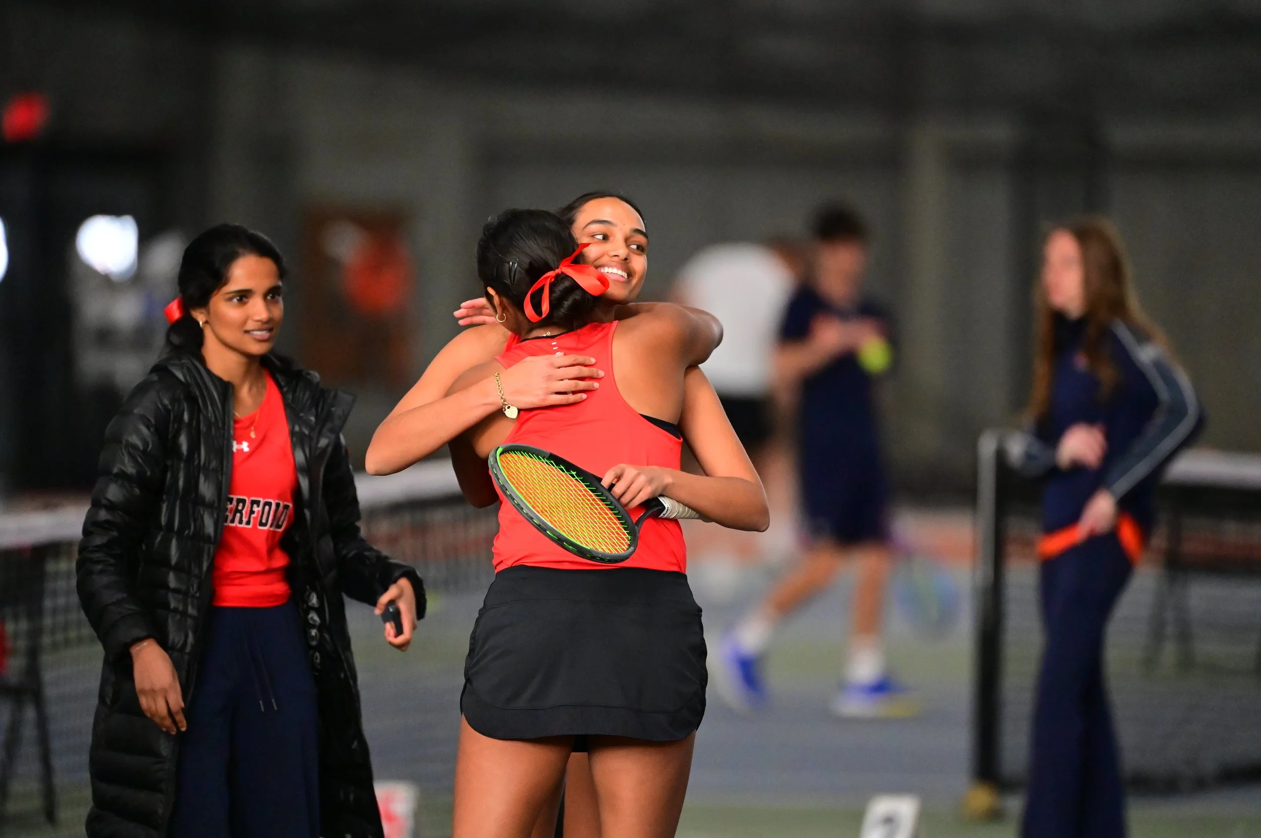 Two female tennis players hugging on an indoor tennis court, with other players and a referee in the background.