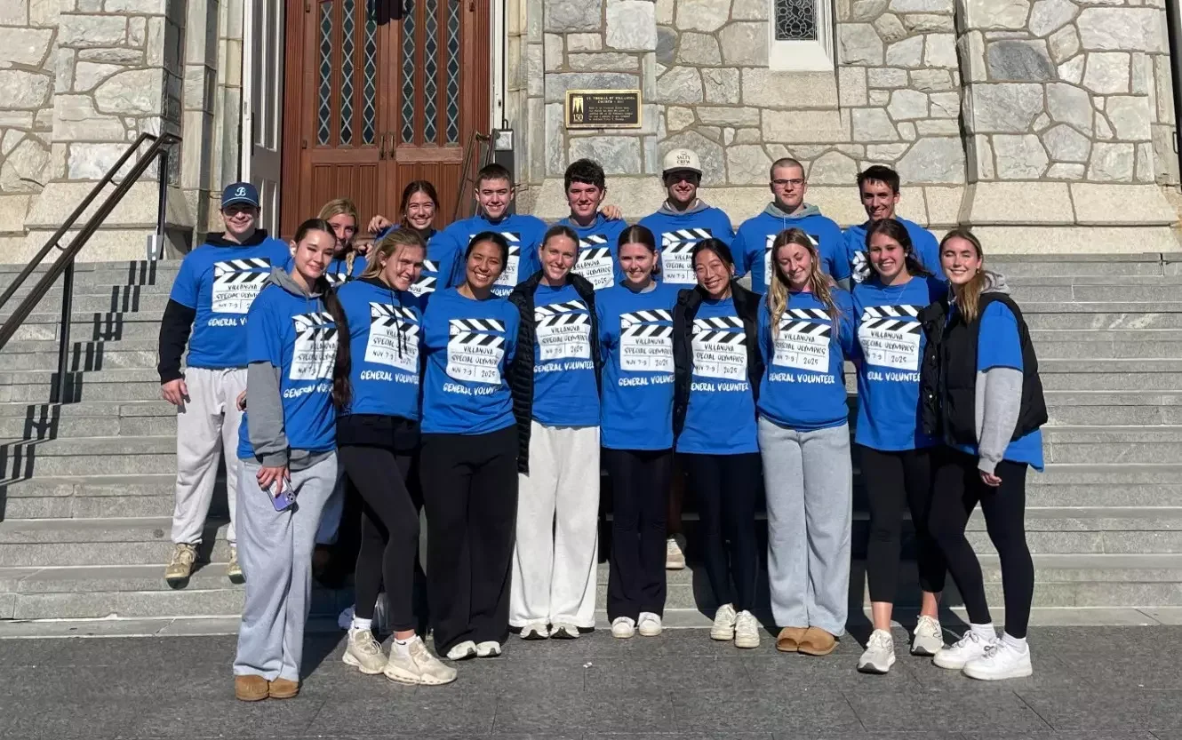 A group of teenagers standing on the steps outside a stone building, wearing matching blue t-shirts that say 'Villanueva Special Olympics Nov 7-9 2023', smiling for a group photo.