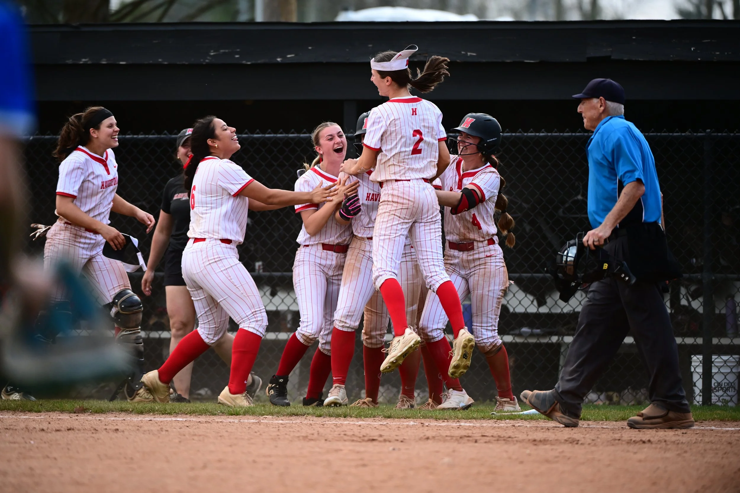 The Haverford College softball team celebrating a victory on Class of '95 Field, with one student-athlete being lifted by teammates, while a male umpire stands nearby smiling.