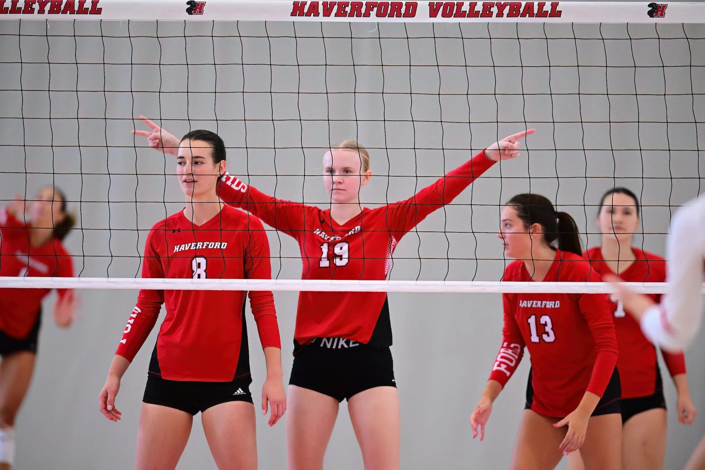 Haverford Volleyball players in red uniforms on the court, with a net and a banner reading 'Haverford Volleyball' overhead.
