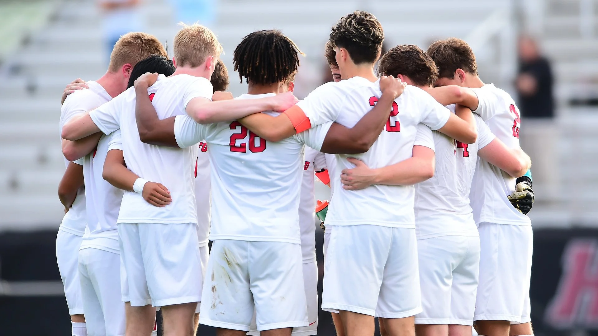 Group of Haverford Men's Soccer soccer players dressed in white uniform kits with red lettering share a pre-match huddle.