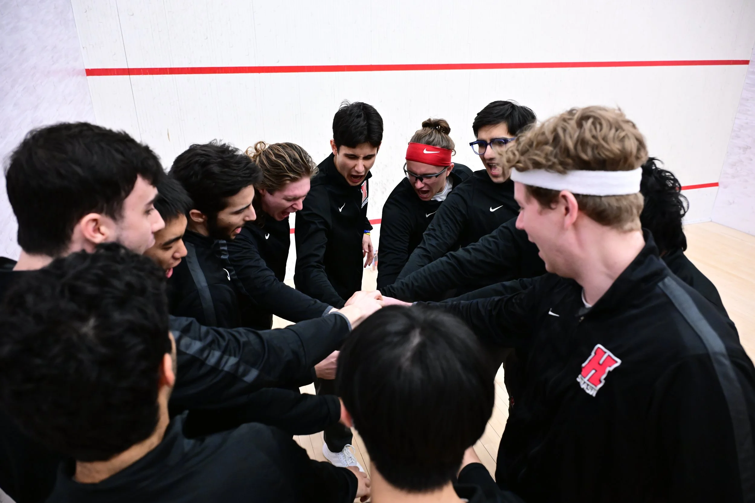 Haverford Men's Squash team dressed in black and red uniforms huddled together in a circle before a match.