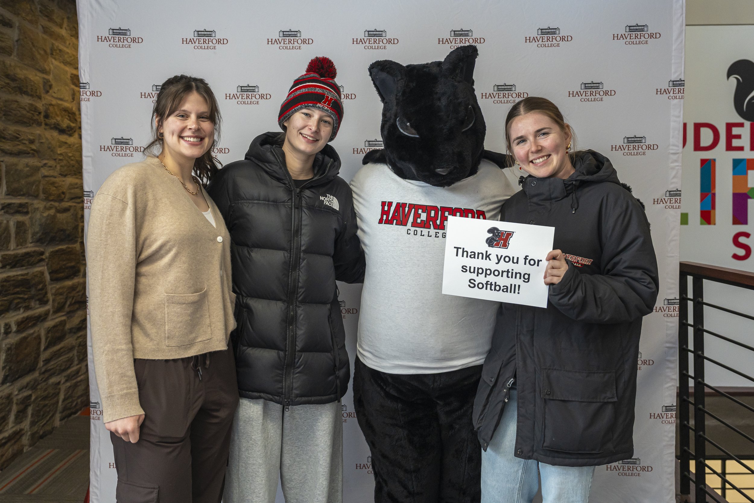 Four people standing together in front of a backdrop with Haverford College logo. One person is dressed in a full black plush animal costume with a sign that says 'Thank you for supporting Softball!' Another person is wearing a black jacket and holding the sign. The two women on the sides are smiling and wearing casual clothing.