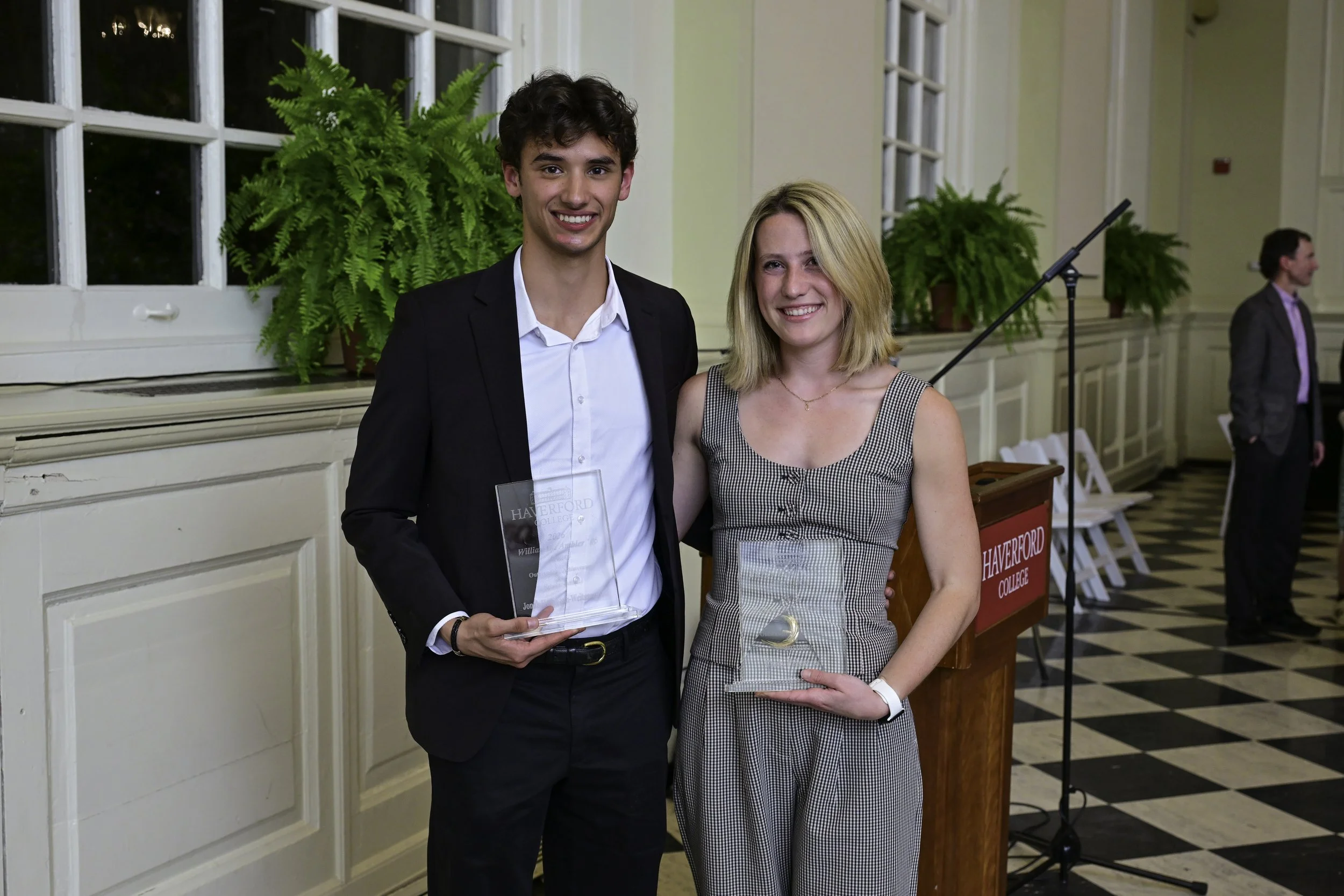 A young man and woman dressed in formal attire are standing together indoors, smiling at the camera. The man is holding a clear award, and there are large green plants and other people in the background.