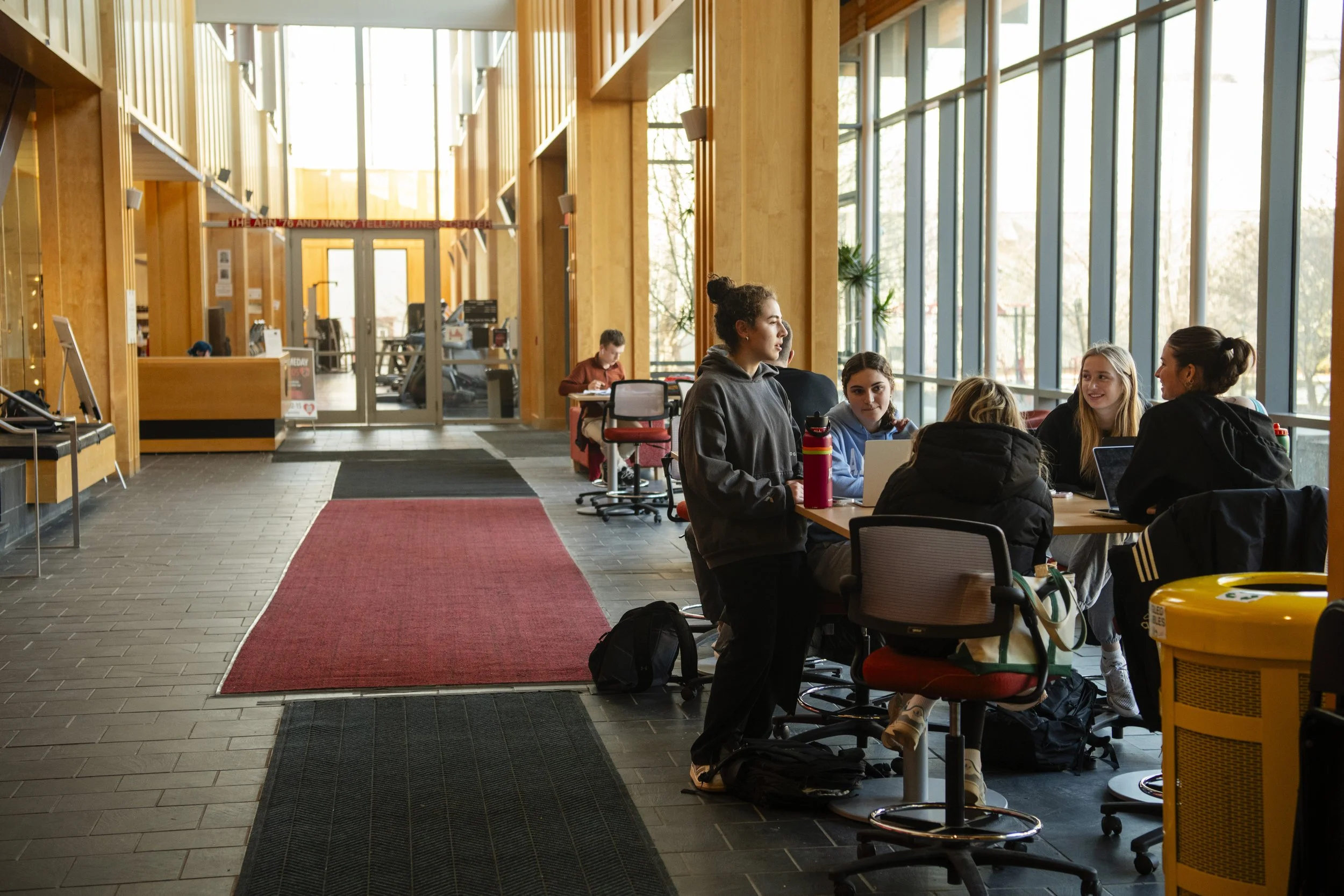 People sitting at tables in a spacious, well-lit indoor space with large windows and wooden accents, possibly a lounge or cafeteria.