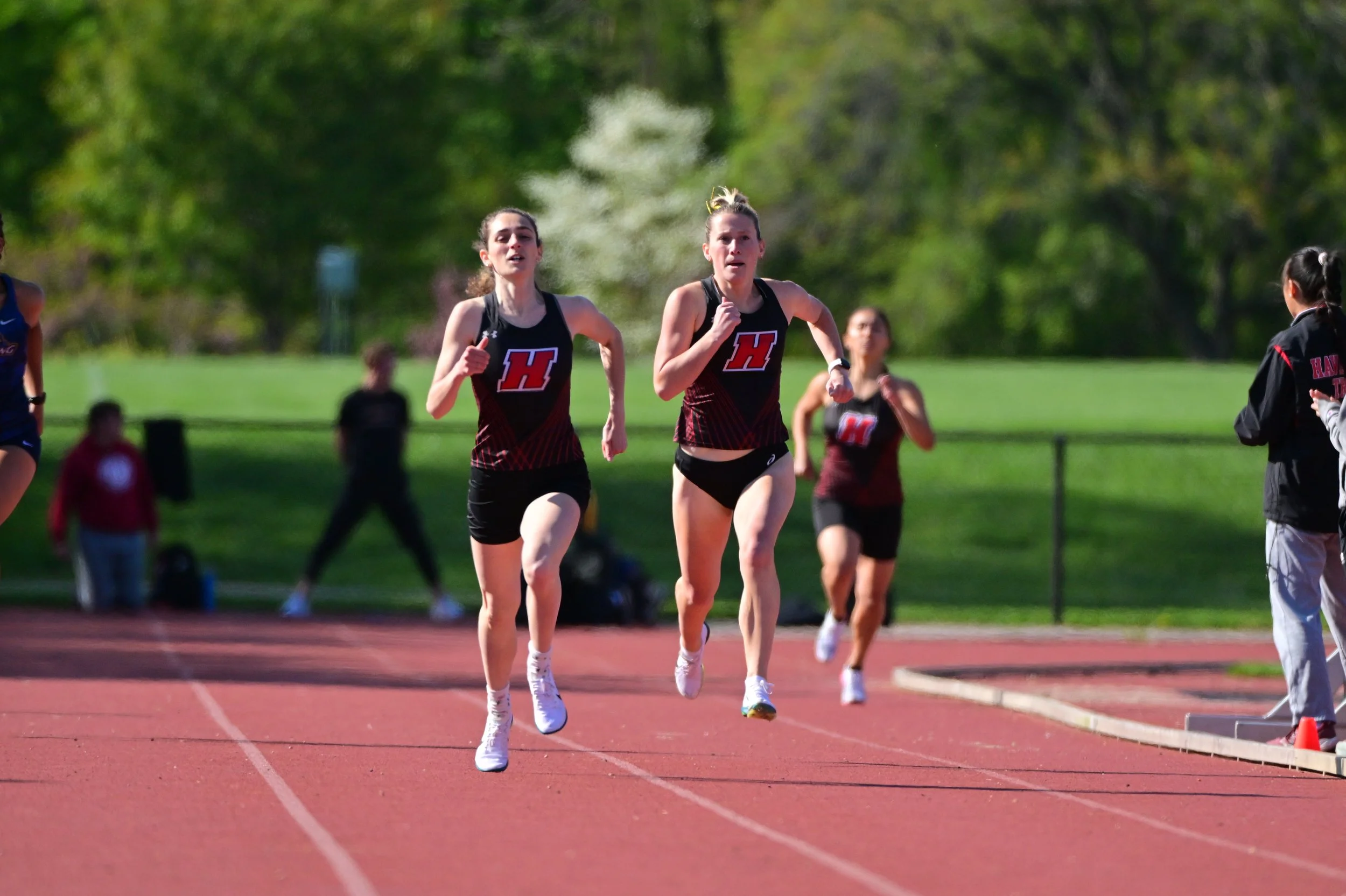 Two female athletes running on a track during a race, wearing black and red uniforms with a large red letter 'H' on their shirts, with green trees and other runners in the background.