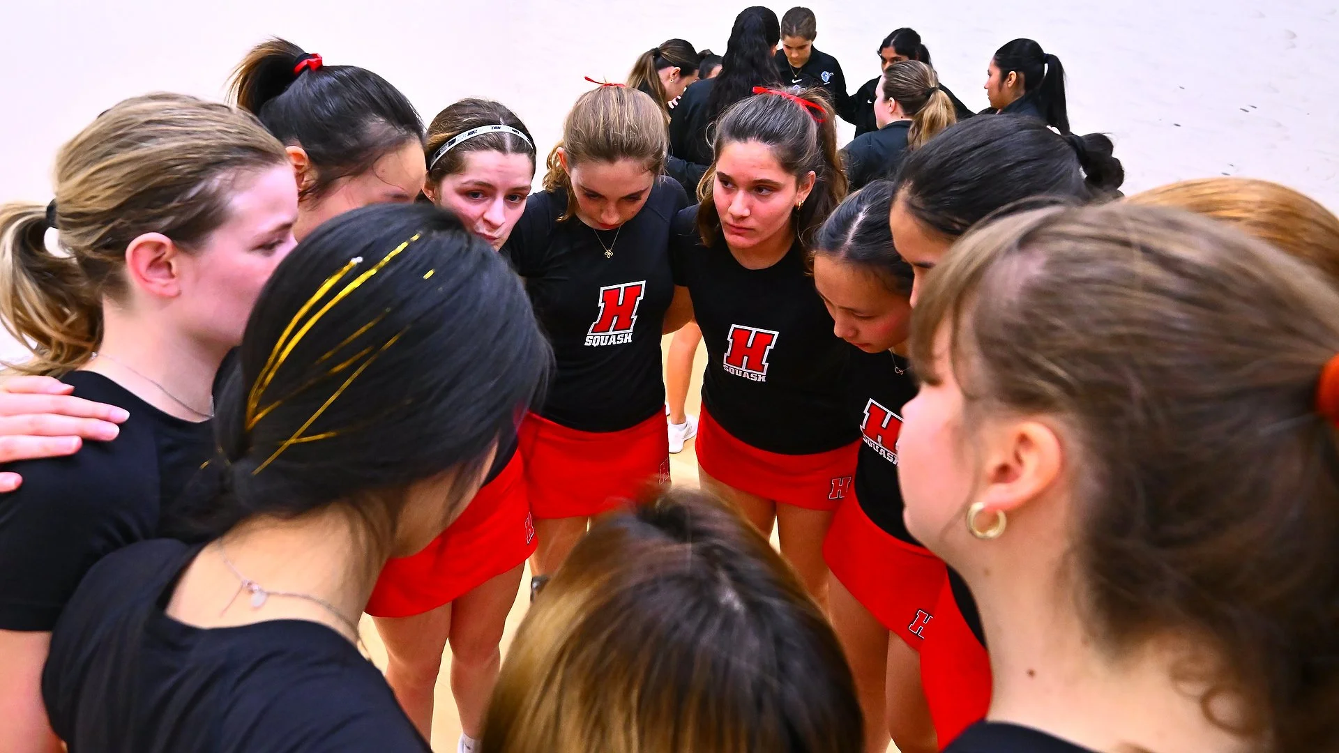 Haverford Women's Squash team dressed in black and red uniforms huddled together in a circle before a match.