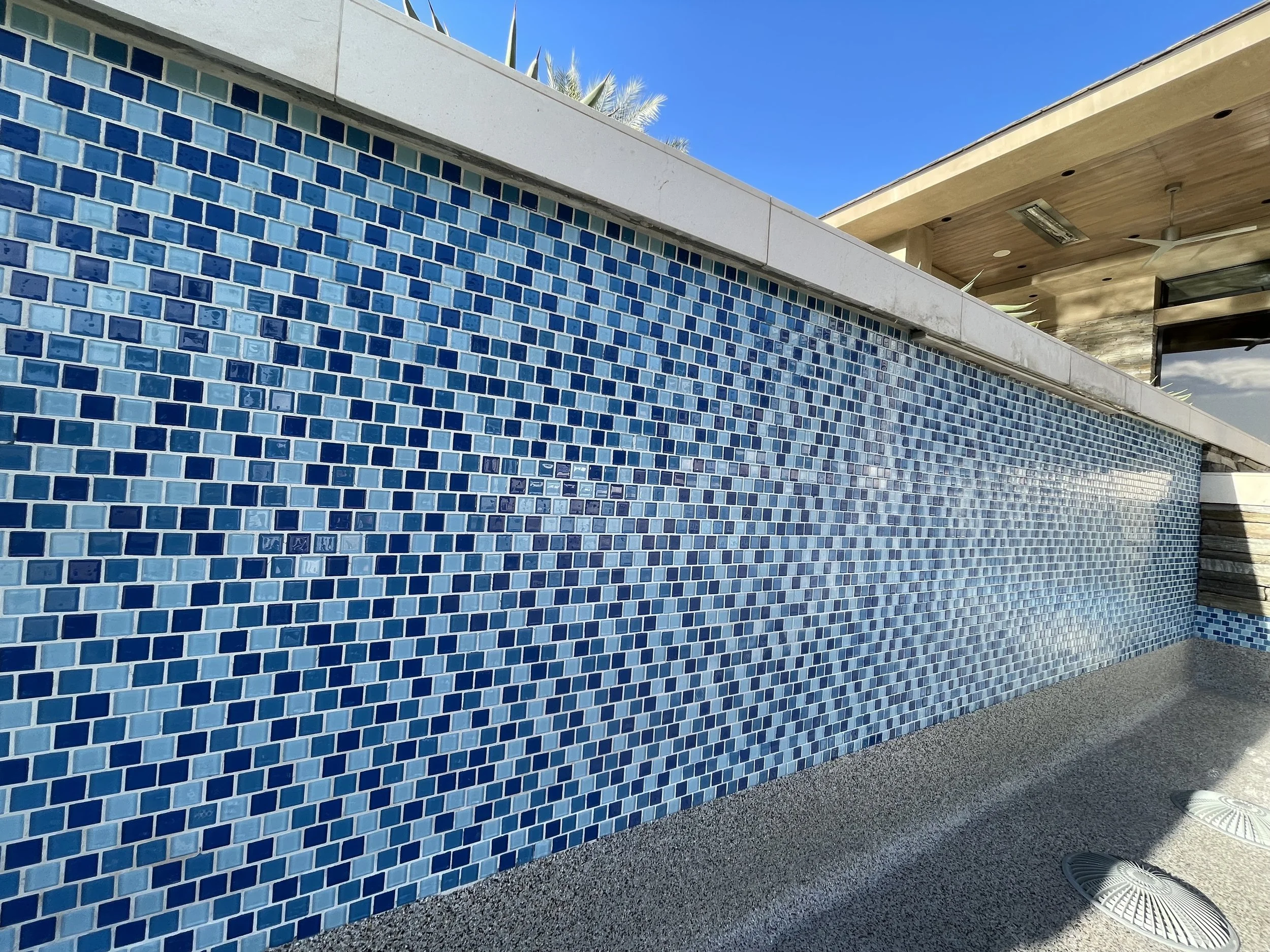 A long outdoor swimming pool with blue mosaic tile walls, a concrete edge, and a covered seating area with ceiling fans under a wooden roof.
