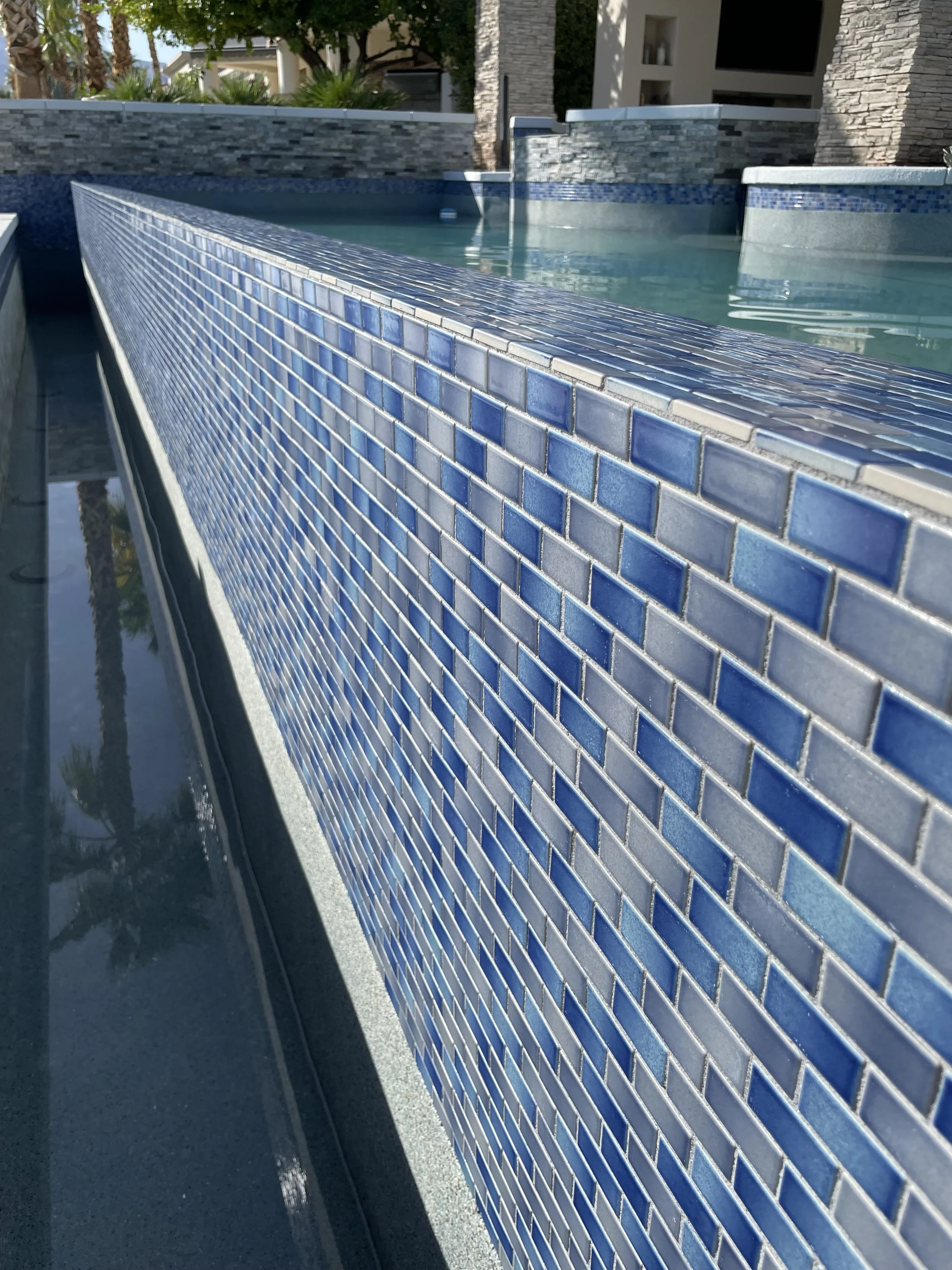 Close-up of a blue-tiled ledge alongside a swimming pool with a modern house in the background.