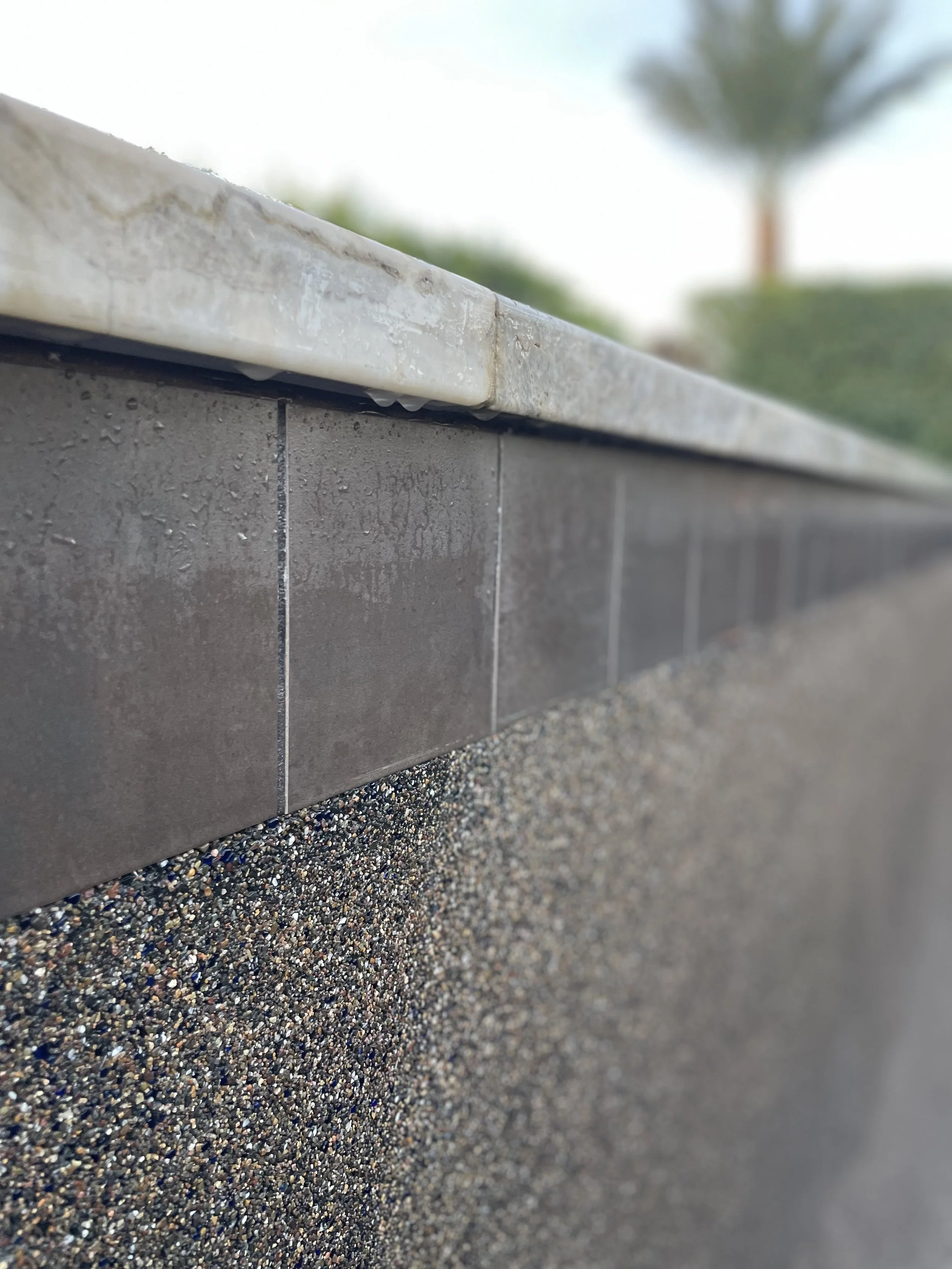 Close-up of a textured concrete wall with a smooth, brown tile border and a light-colored marble cap on top, with blurred trees in the background.