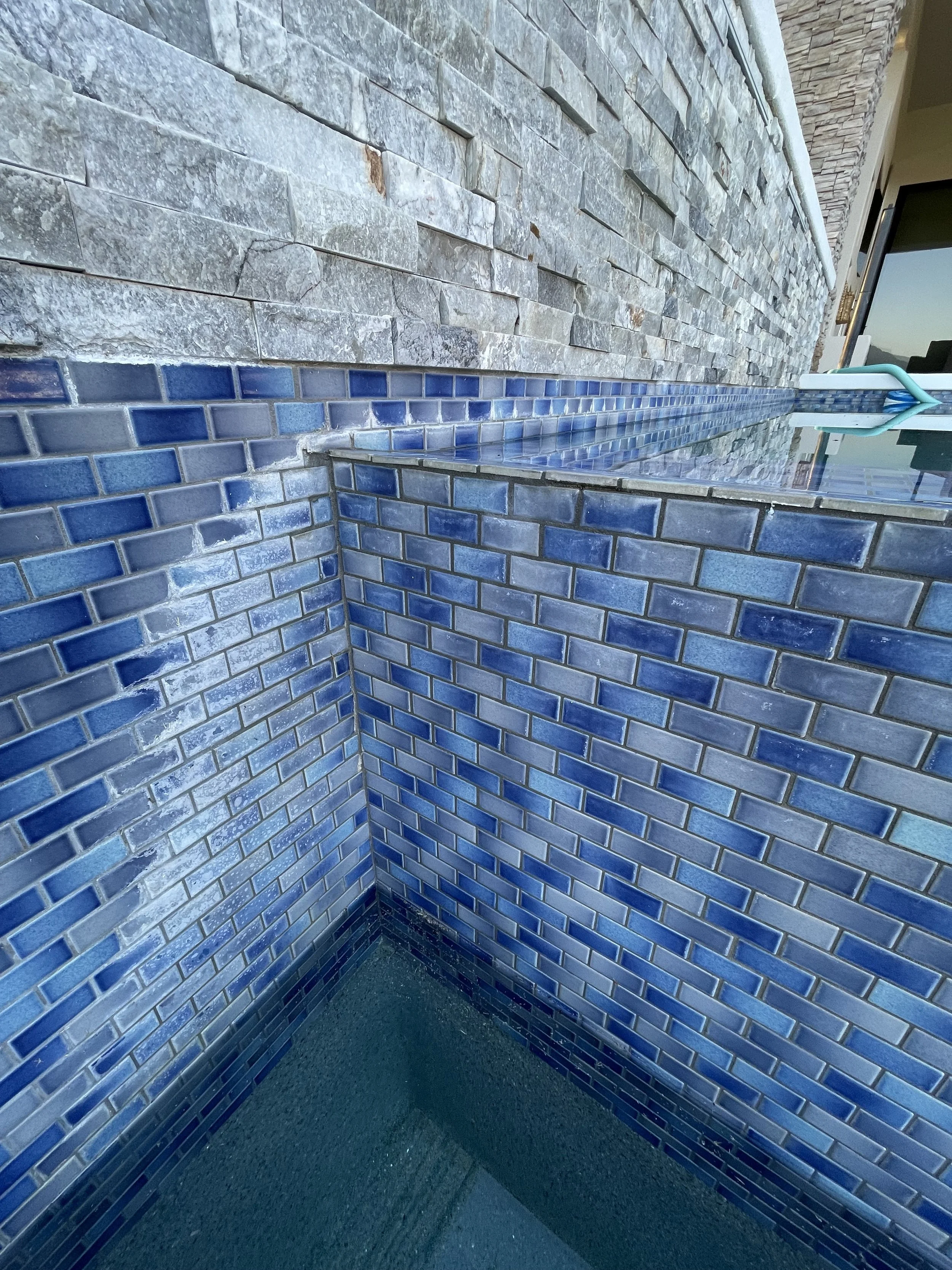 Close-up of a swimming pool with blue tile interior and a water hose at the edge, overlooking a stone wall and outdoor area.