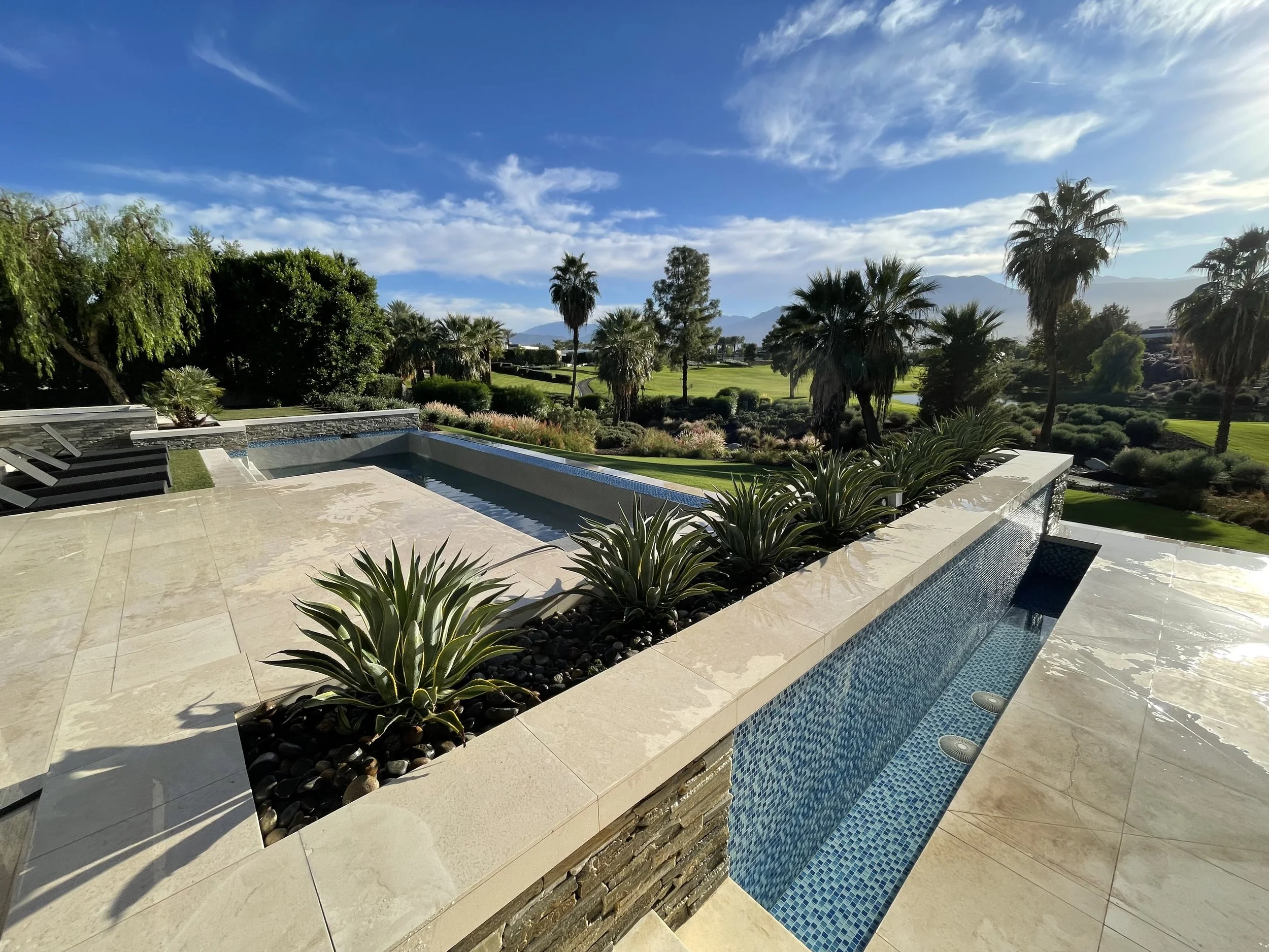 Modern backyard with a rectangular infinity pool, desert plants, and lush greenery, including tall palm trees, overlooking a golf course under a bright blue sky with some clouds.