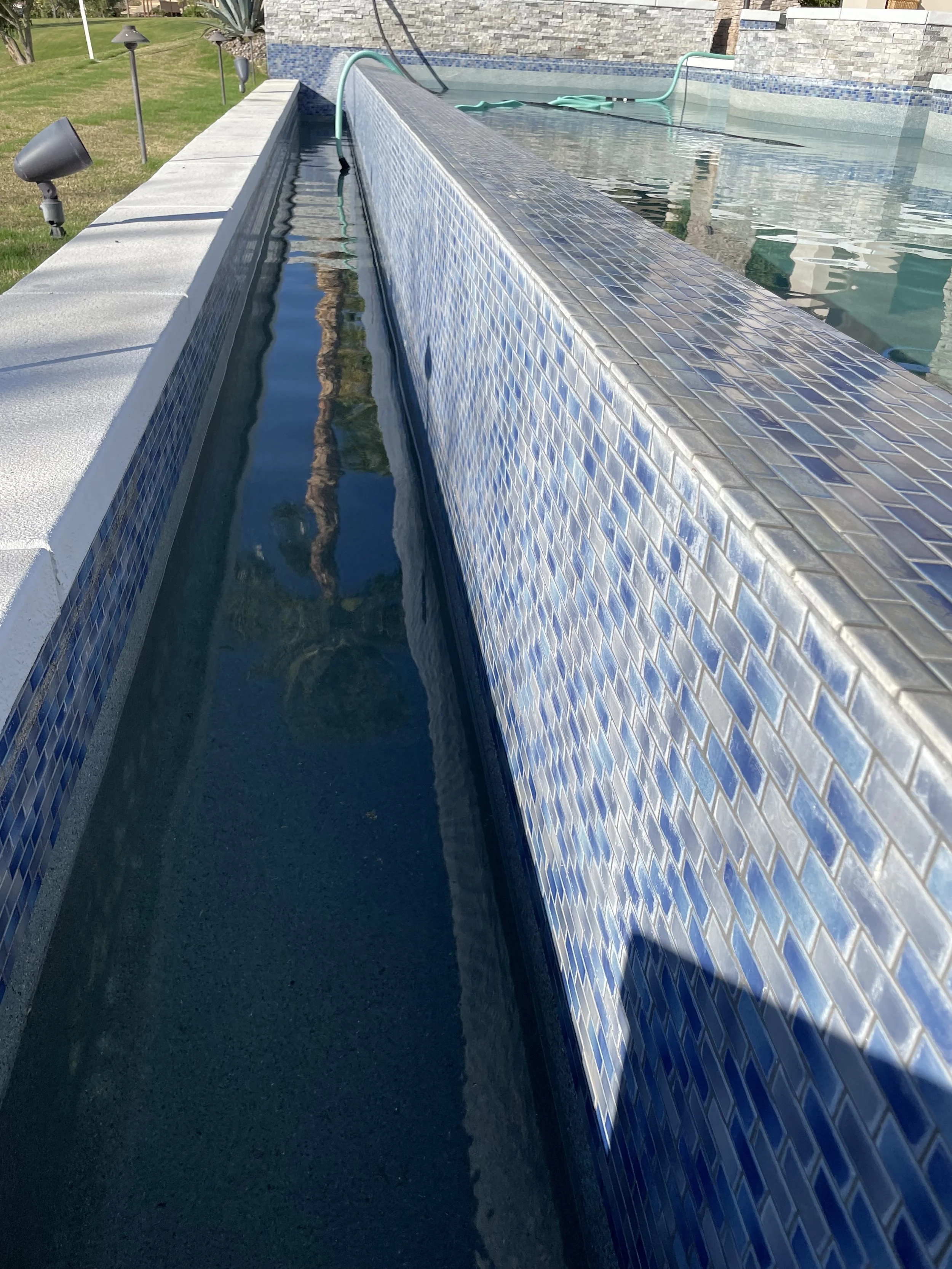 Close-up view of a swimming pool with blue tile lining, showing the water level and a pool cleaning hose in the background. The pool is outdoors with sunlight reflecting on the water and surrounding area.