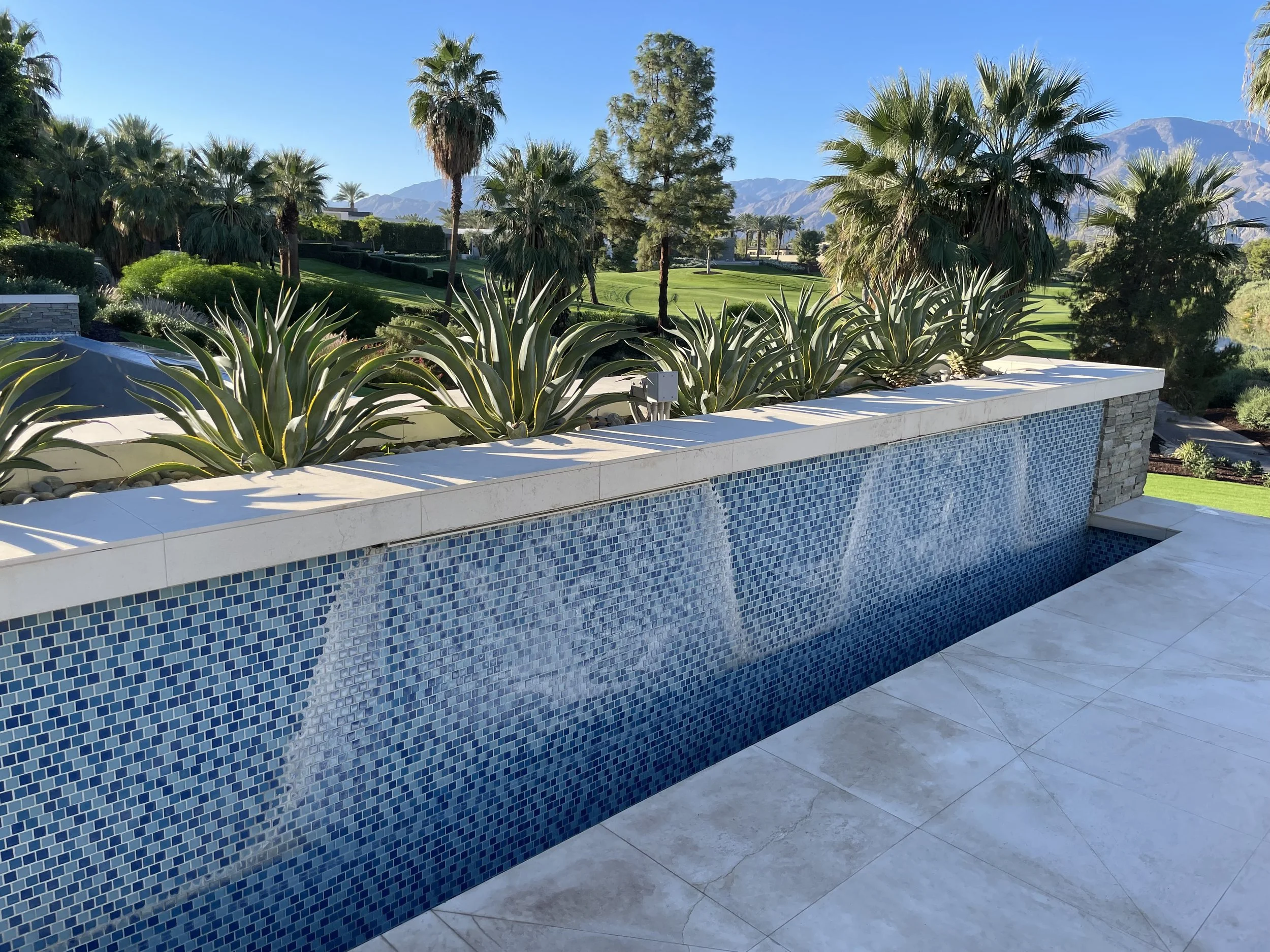 A modern outdoor water feature with cascading waterfalls over a blue mosaic tile wall, surrounded by desert plants including agave, with a lush grassy landscape, palm trees, and mountain range in the background.