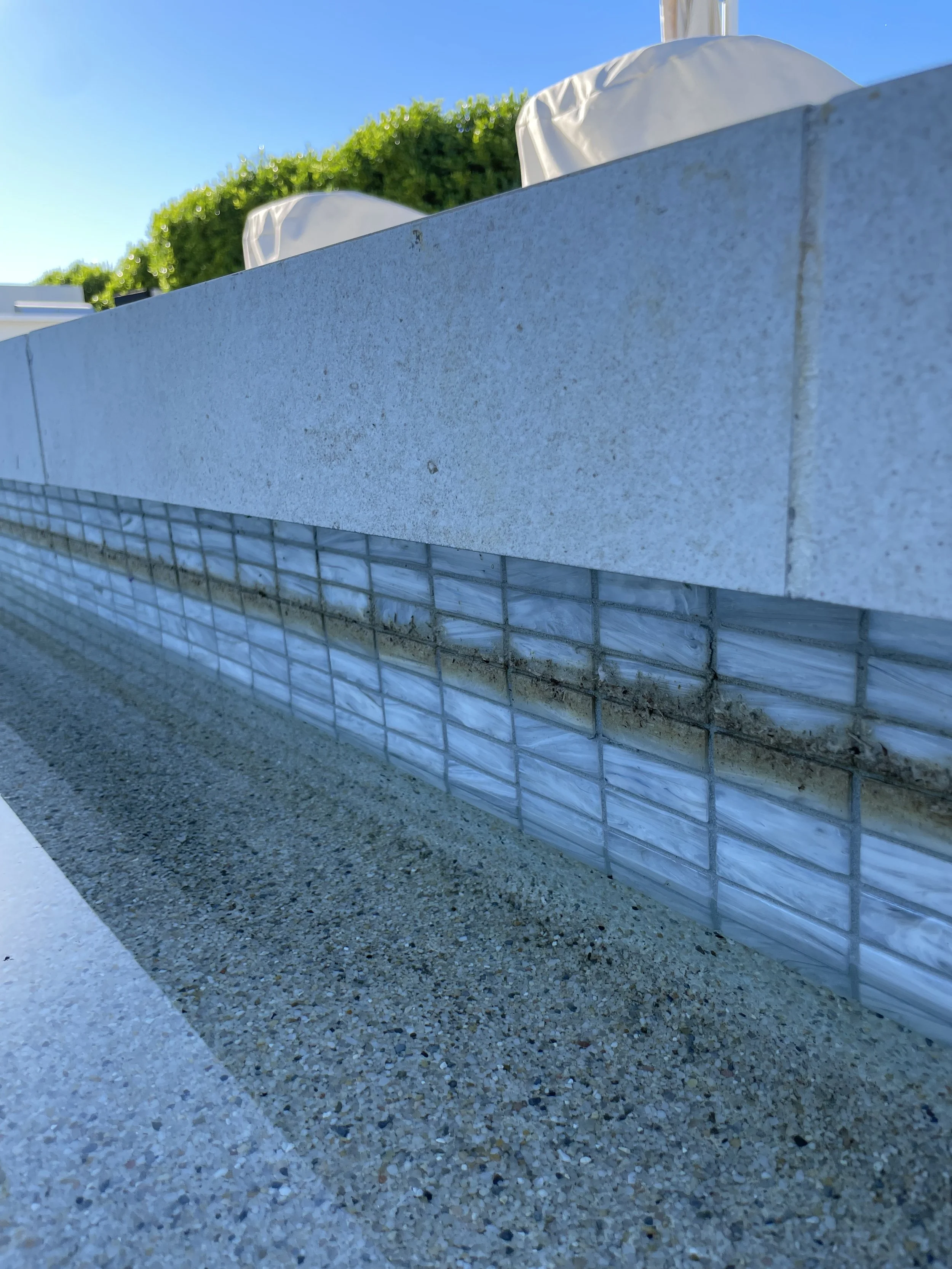 Close-up of a section of a building exterior showing a white tiled ledge with grout lines, a wall with light-colored bricks, and gravel ground underneath, with greenery and blue sky in the background.