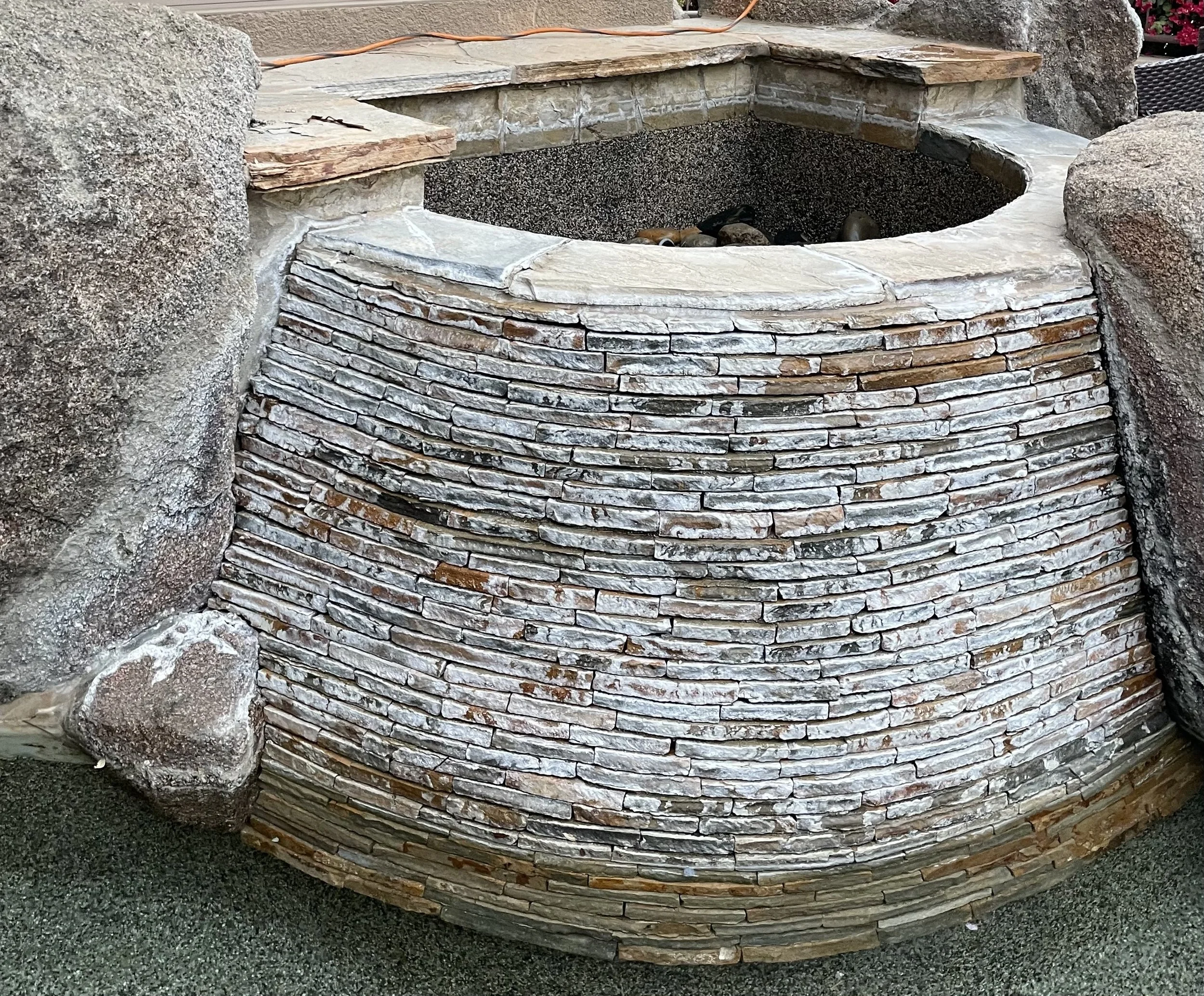 A half barrel shaped outdoor stone water fountain with a flat top, surrounded by large rocks and stones, and a dark inside basin filled with rocks.