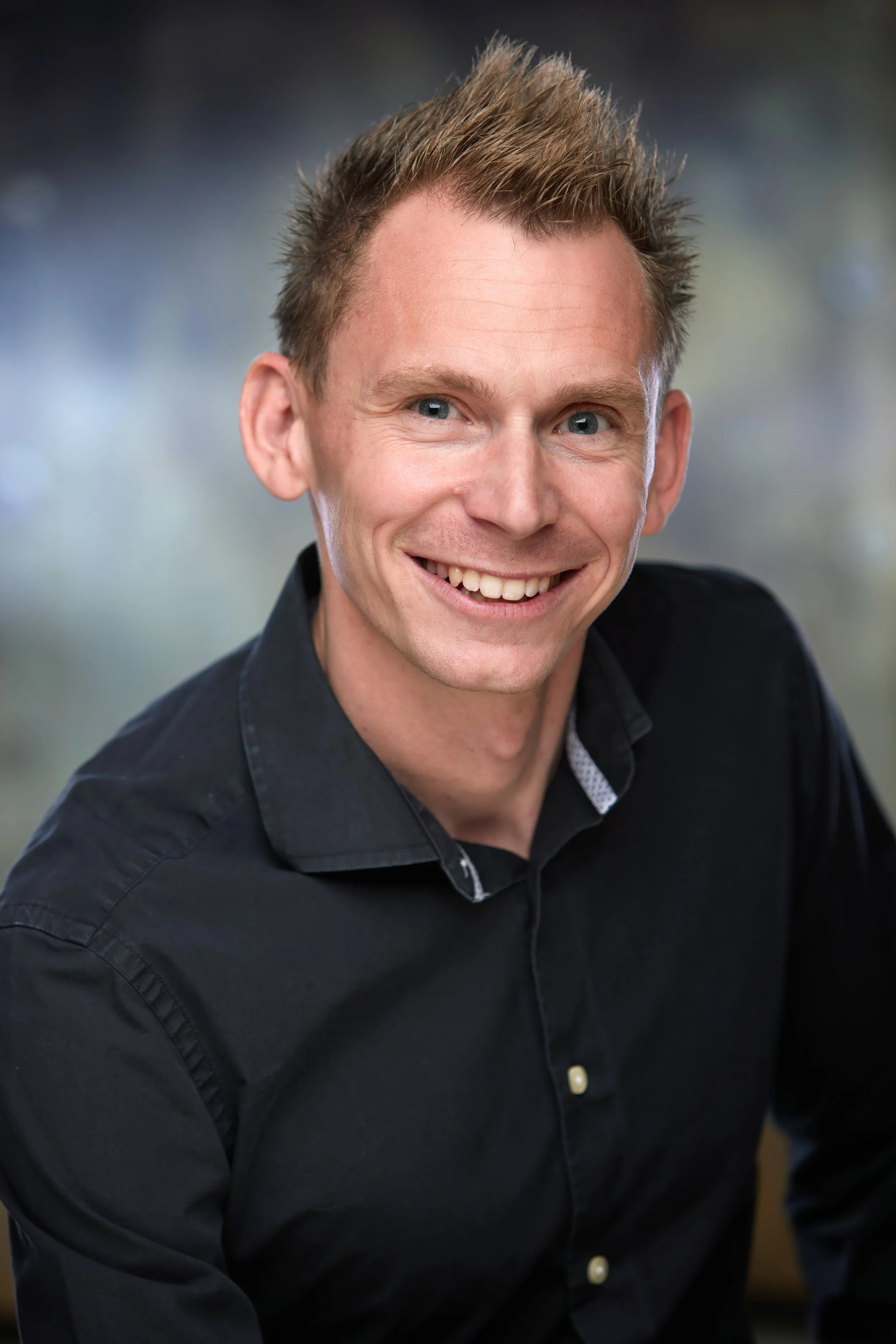 A smiling man with short, light brown hair, wearing a black button-up shirt, photographed against a blurred background.