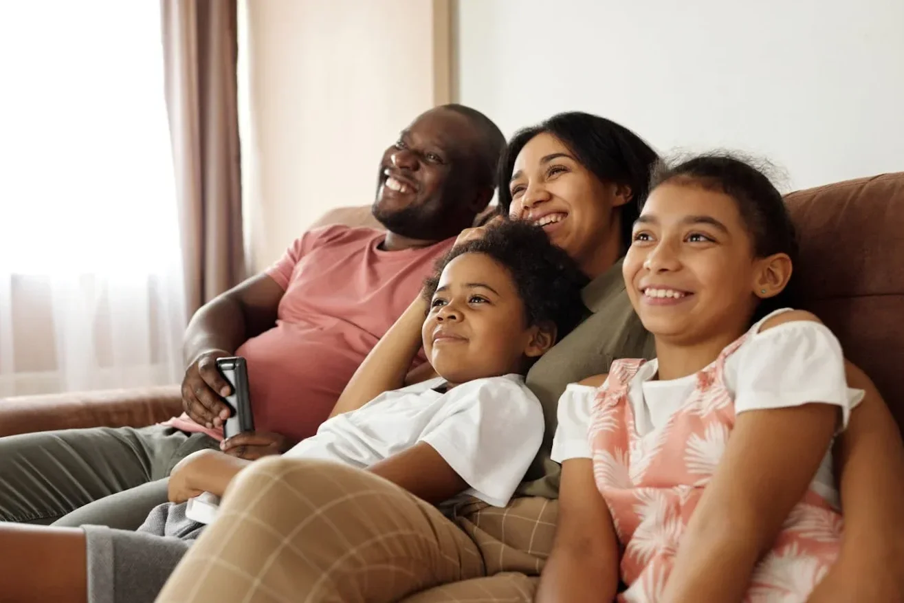 Family of four relaxing on a brown couch, smiling and enjoying time together in a living room with curtains and natural light.