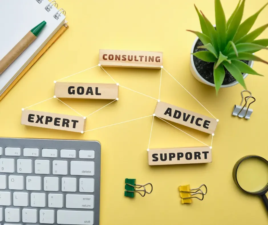 A yellow desk with a keyboard, notepad with a pen, a potted plant, binder clips, a magnifying glass, and wooden blocks with words like 'Goal', 'Consulting', 'Expert', 'Advice', and 'Support' connected by strings, representing consulting or business advice concepts.