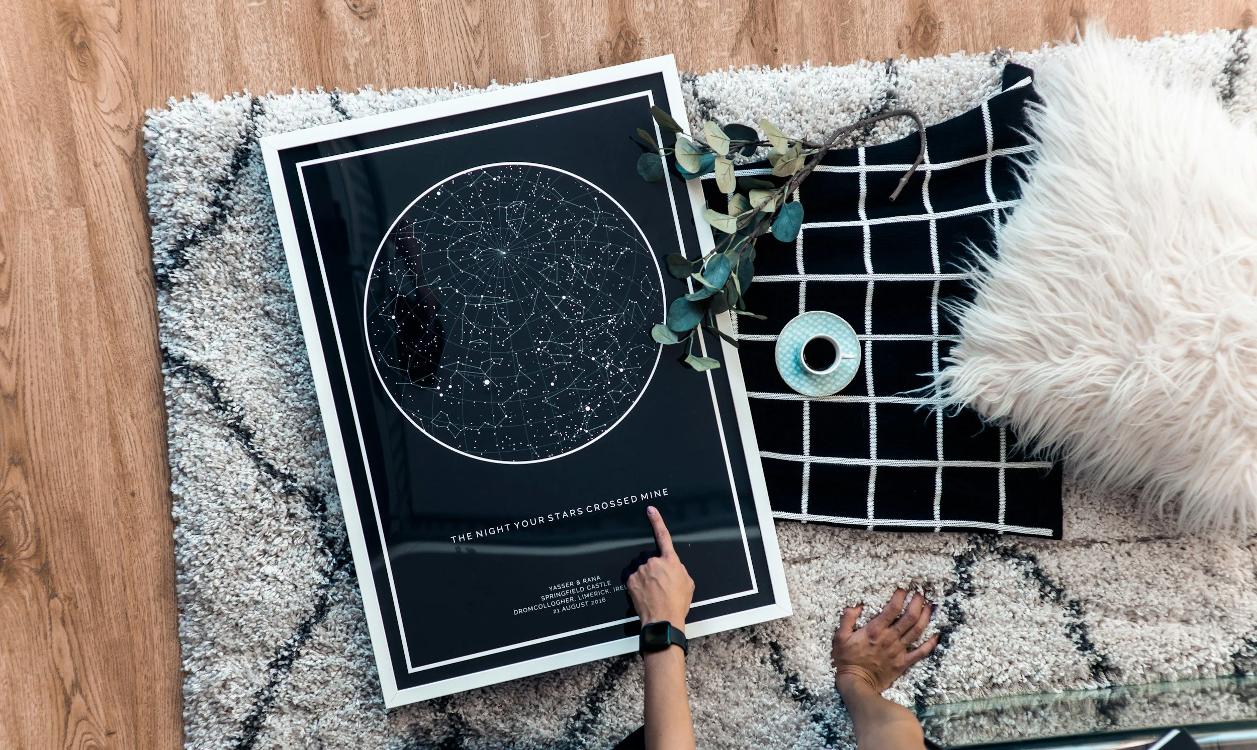 A poster of a star constellation map with a person's hand pointing at it, a small cup of coffee, and a sisal pillow on a black and white checkered cloth on a fuzzy rug.