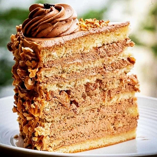 A close-up of a slice of layered caramel cake with chocolate frosting and nuts on the sides, topped with chocolate frosting and a swirl of frosting on top.
