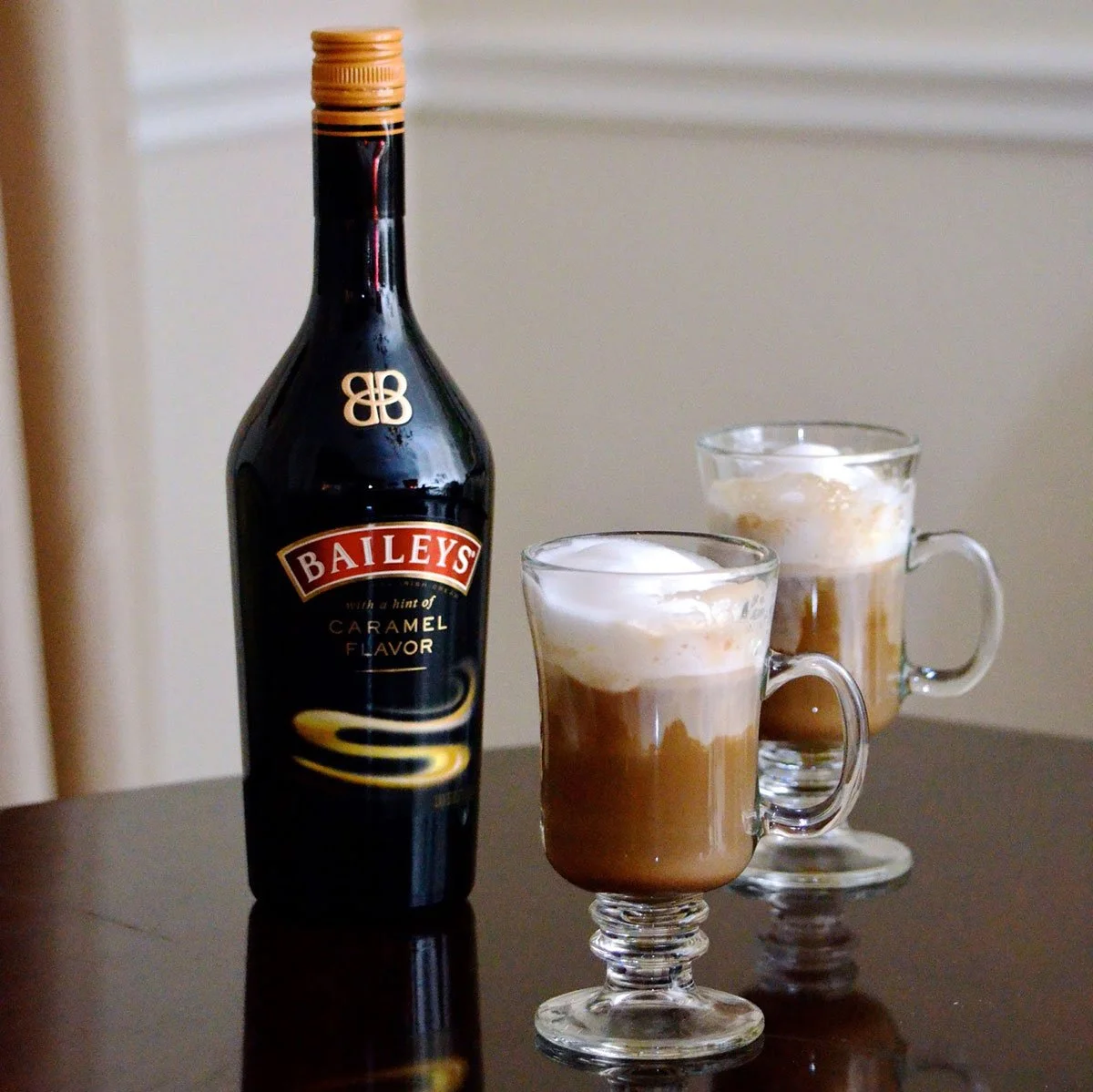 A bottle of Bailey's caramel flavored Irish cream liqueur and two glass mugs filled with a layered coffee drink topped with whipped cream on a dark table.