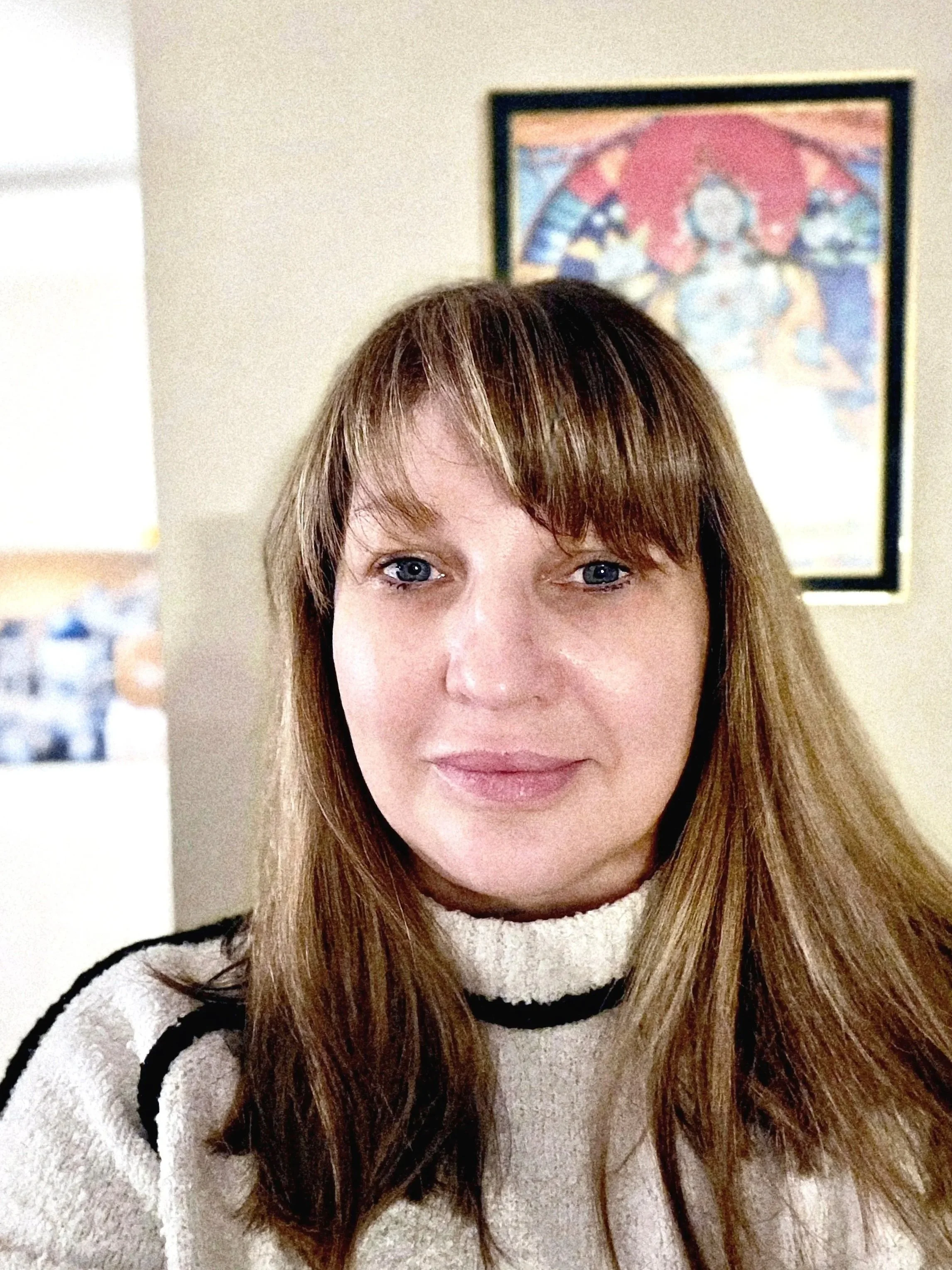 A woman with straight, shoulder-length brown hair and blue eyes, wearing a cream-colored sweater with black stripes, standing indoors with a framed colorful poster in the background.