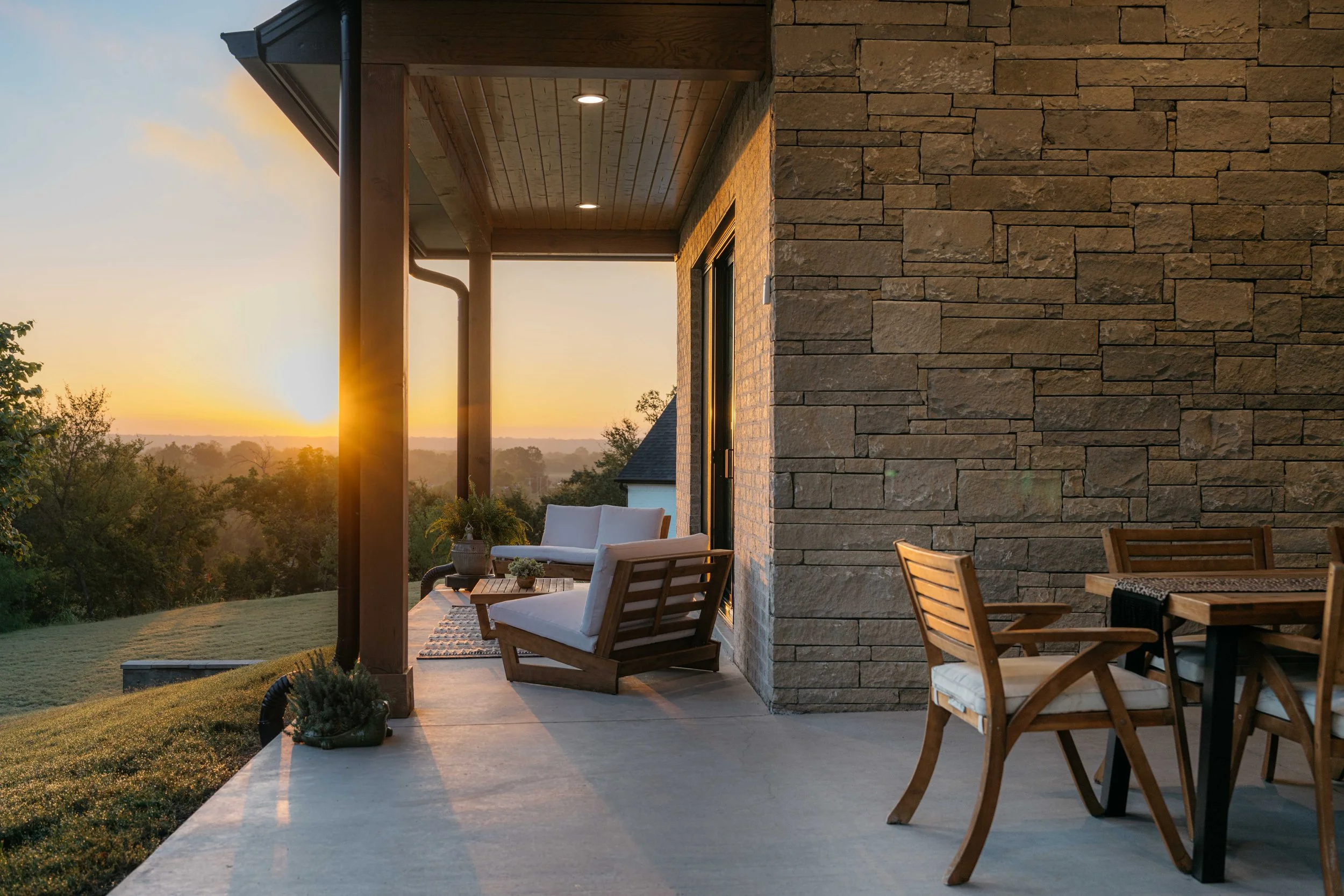 Luxury custom home covered patio with stone exterior, wood ceiling, and outdoor seating overlooking a scenic sunset.