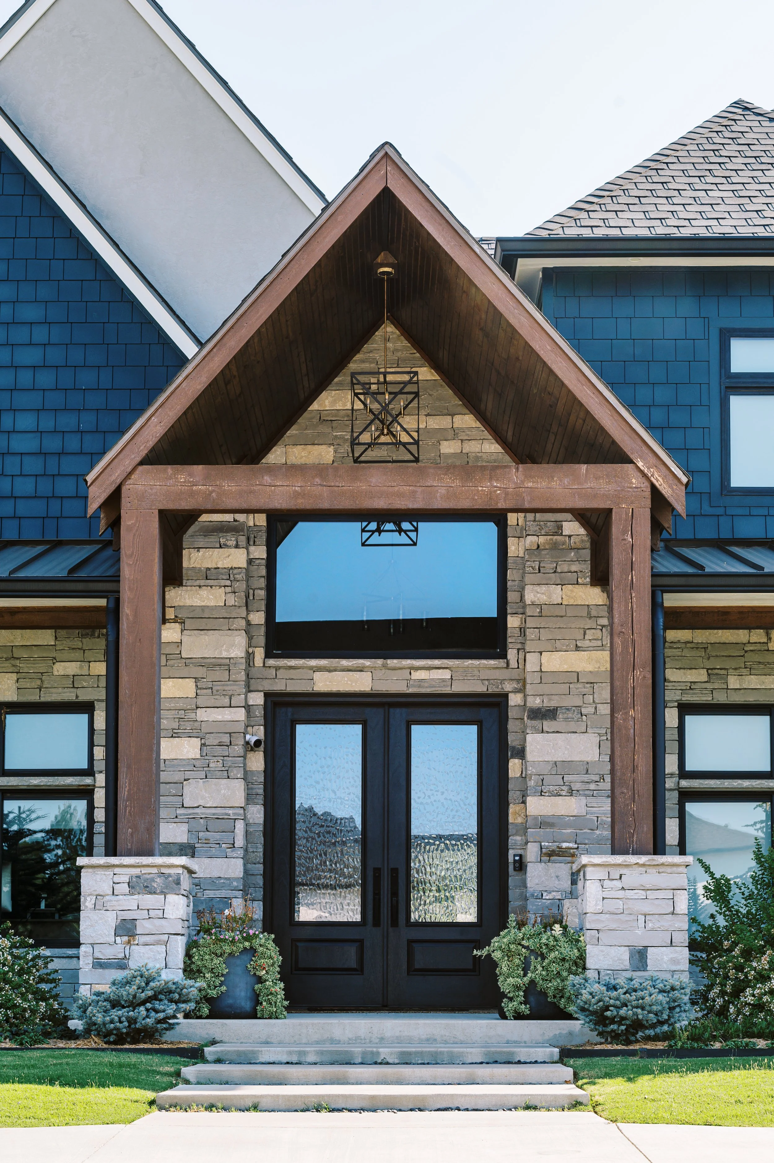 Front entry of a luxury custom mountain home featuring natural stone, timber beams, and modern black double doors, showcasing refined craftsmanship by Snow Peak Customs.
