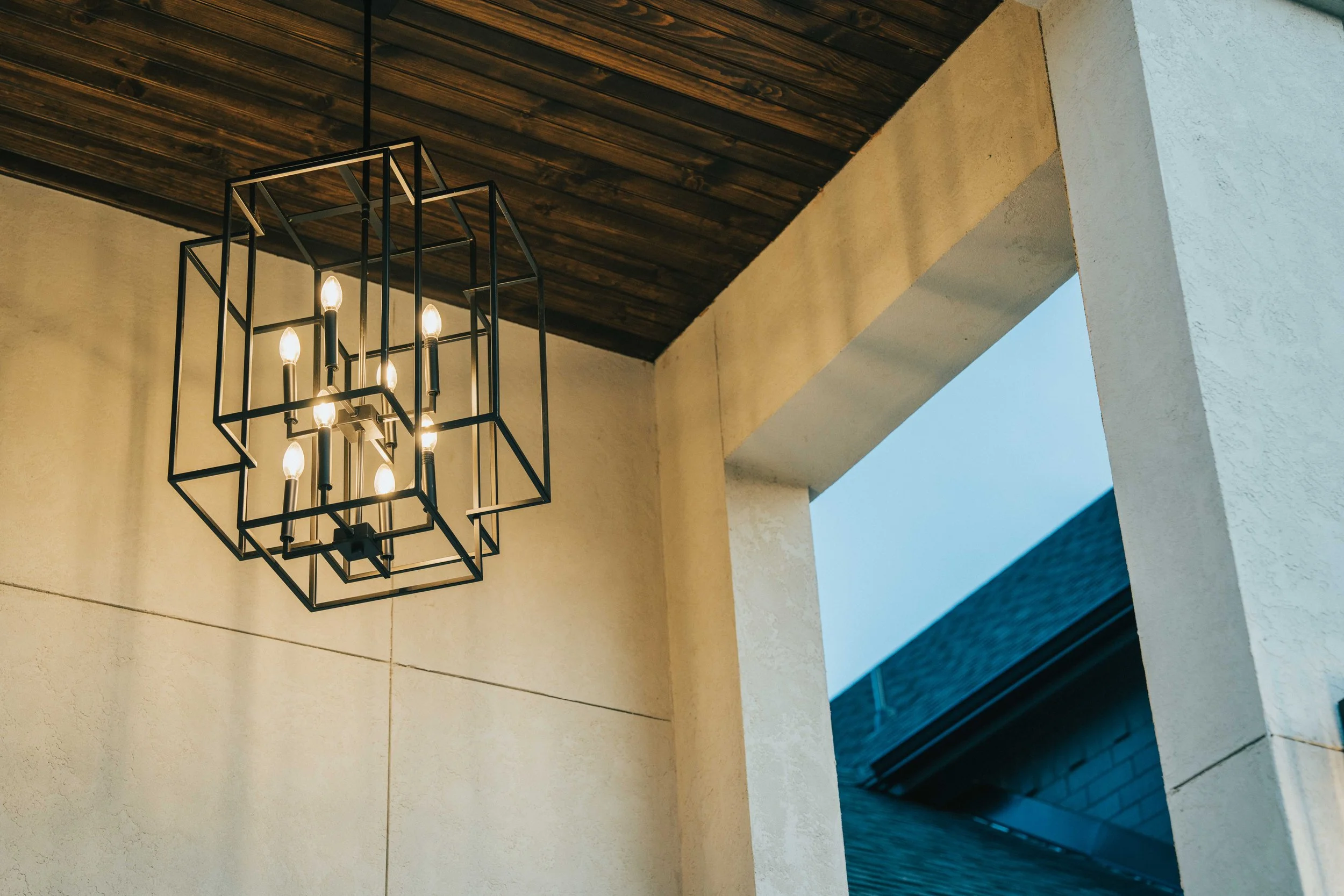 Modern geometric chandelier with black metal frame and warm lighting, suspended from a wood ceiling in a luxury exterior entrance.