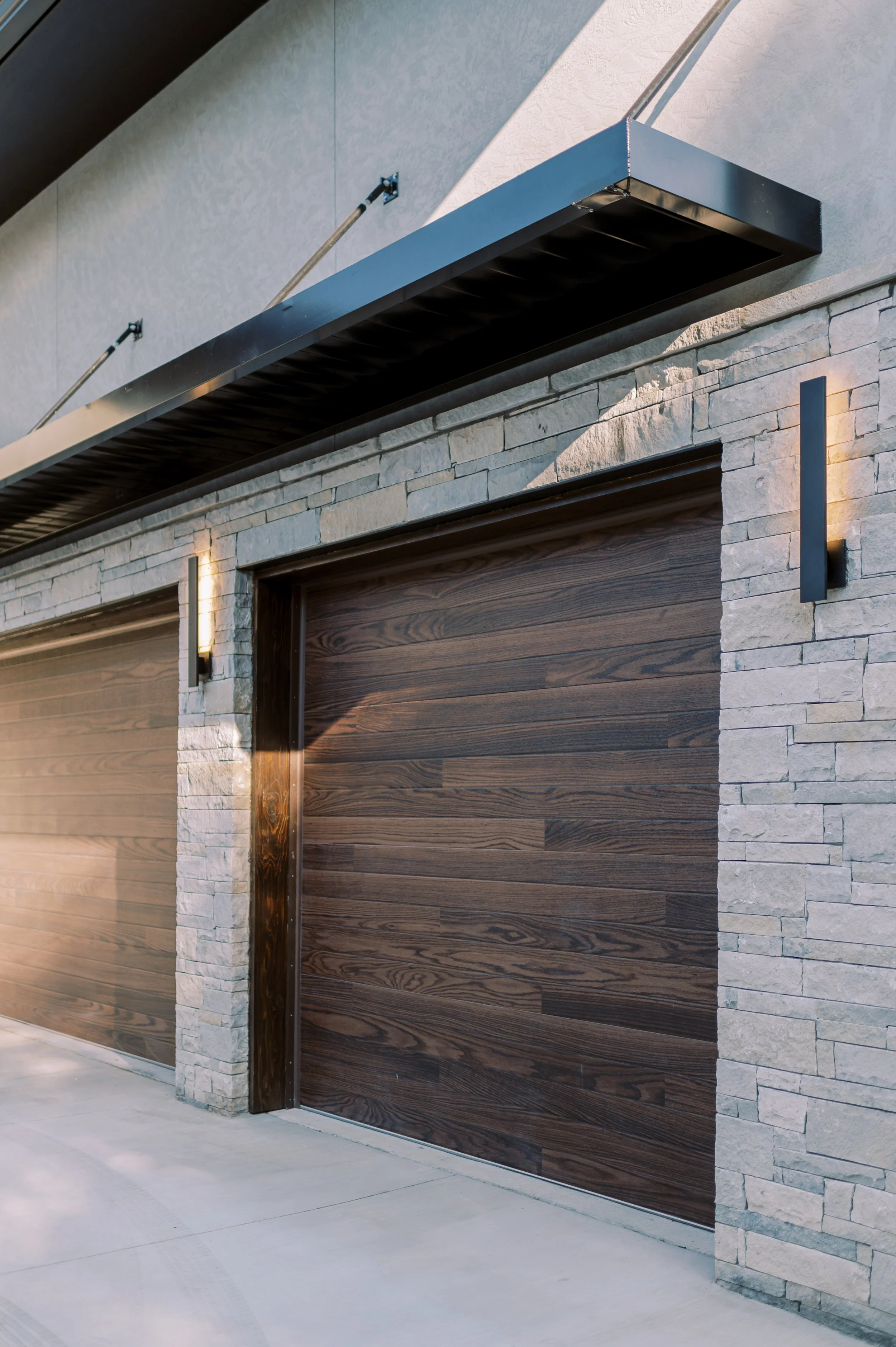 Custom mountain home garage exterior by Snow Peak Customs featuring wood garage doors, stone veneer, and modern metal awning.